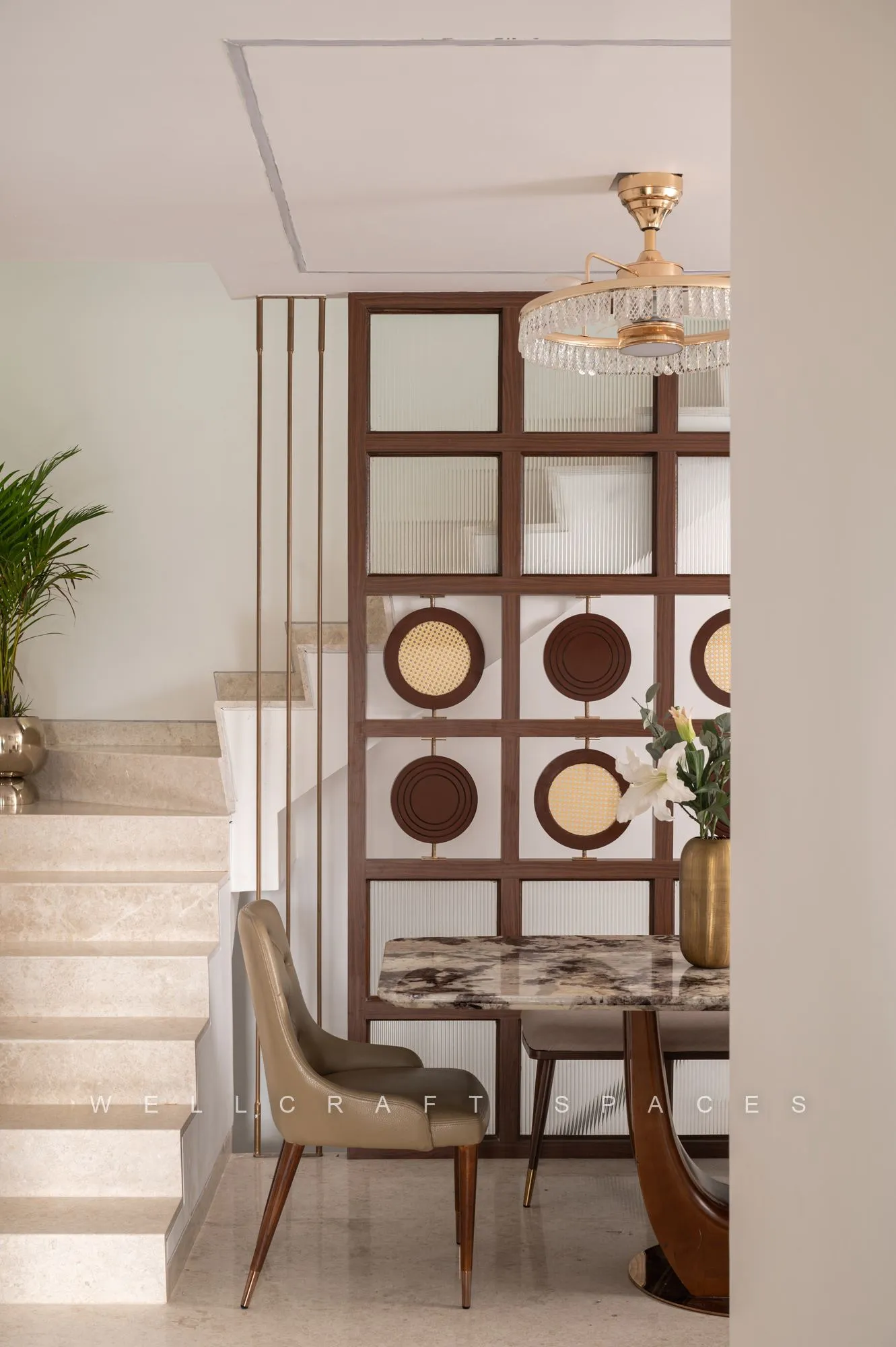 Dining table for four beside the stair, framed by a walnut screen inset with round brass and marble discs, under a tiered crystal pendant.