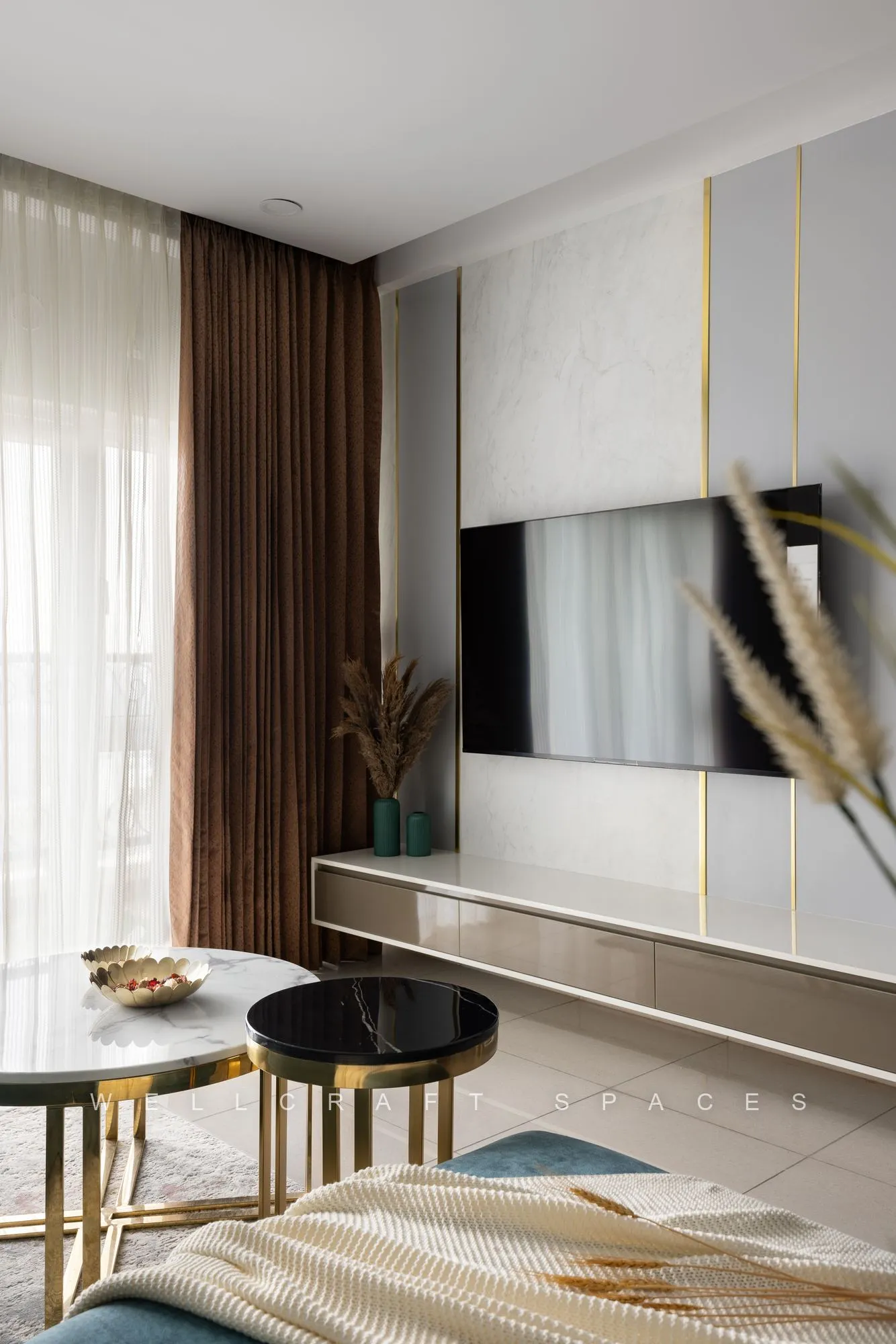 Wall-mounted TV on a cream panelled unit with slim gold inlays, a floating oak console and stacked black-and-gold nesting tables.