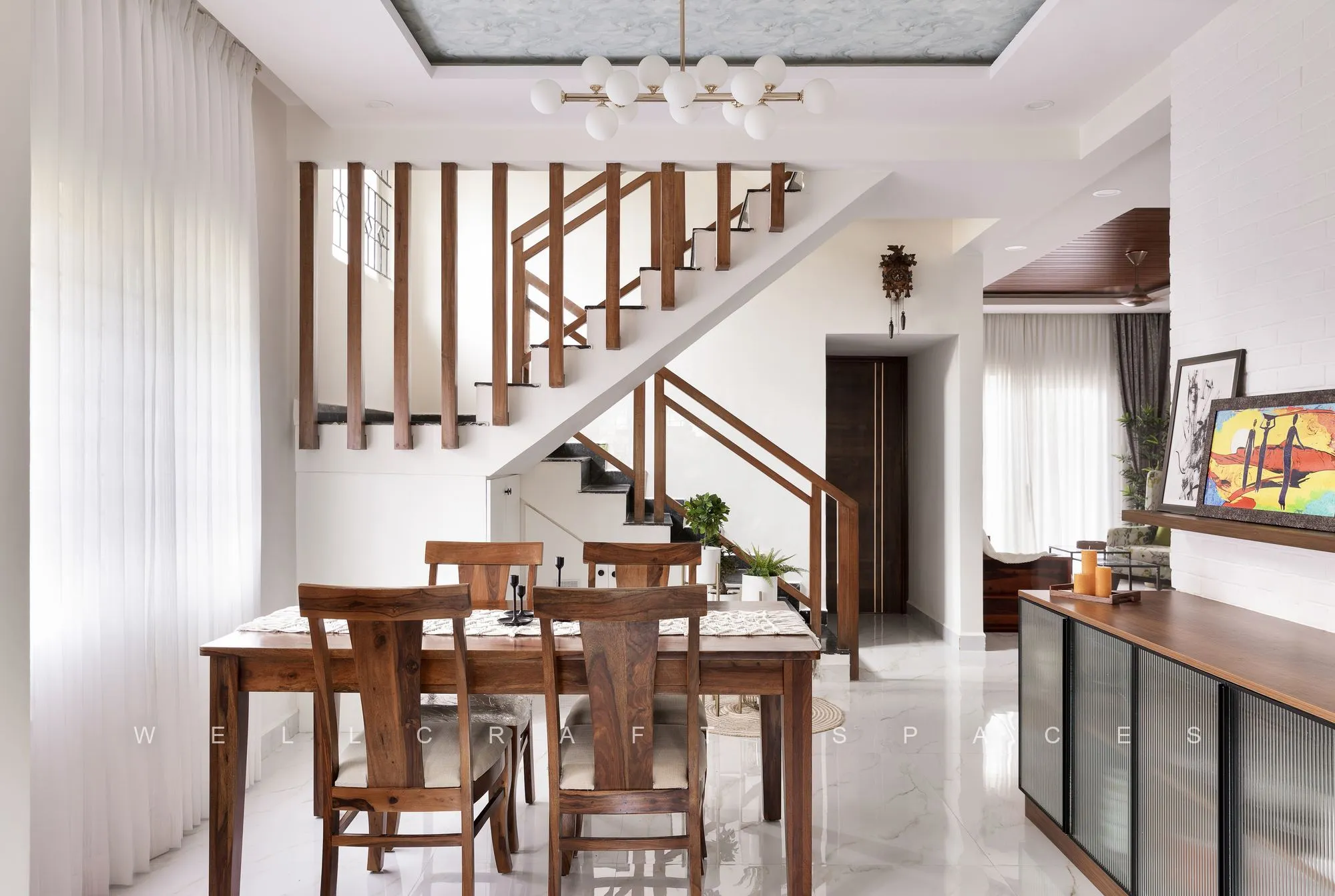Dining room with a solid teak table and cane-back chairs beneath a linear pendant, beside the open timber staircase and a glazed crockery unit.
