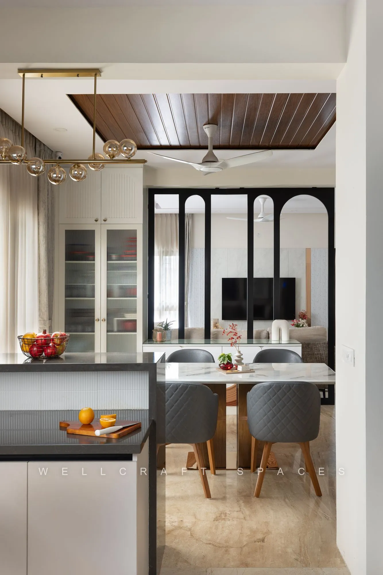 Dining and kitchen seen together, with a grey island counter, glass-fronted crockery unit, slatted wood ceiling and a gold branching chandelier over the table.