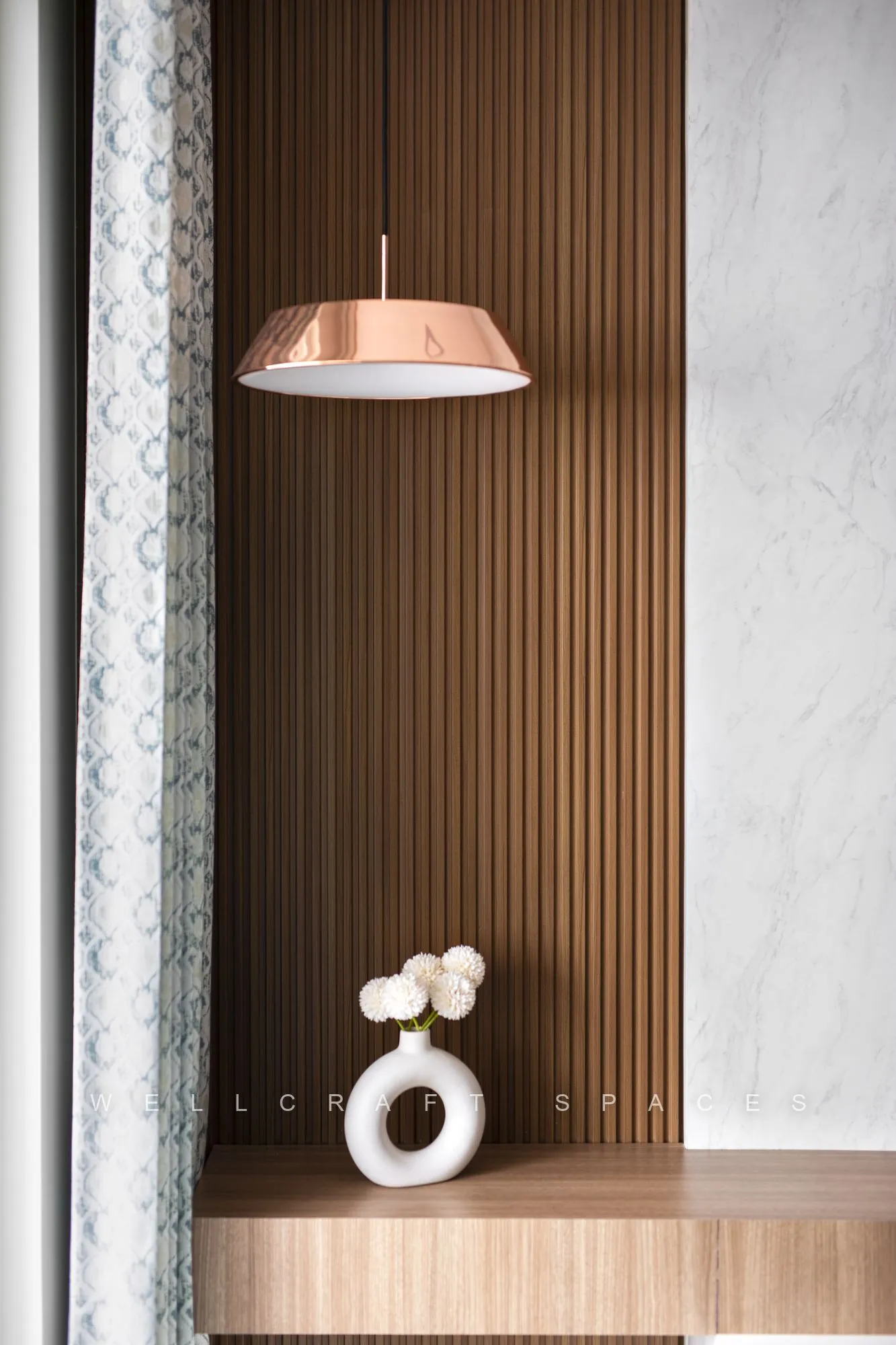 Copper dome pendant over a slatted teak niche styled with a white sculptural ring and a marble-effect wall.