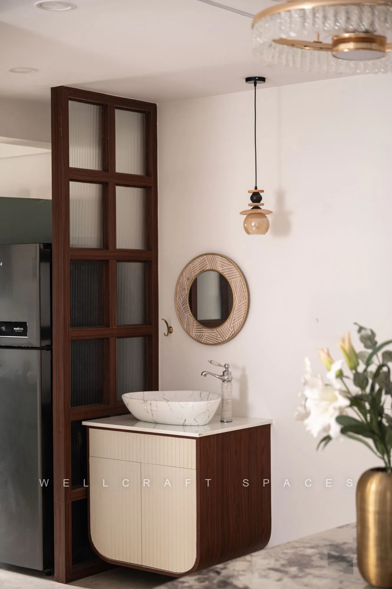 Washbasin counter off the dining room in cream and walnut, with a vessel sink, arched brass mirror and a glass-fronted refrigerator alcove behind.