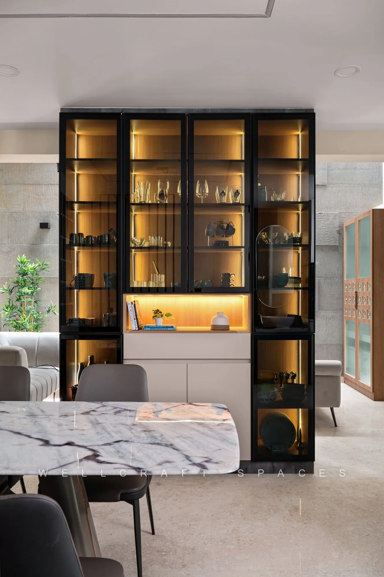 Backlit black-framed crockery unit with glass shutters and a marble counter dividing the dining area, holding glassware and decorative bowls.