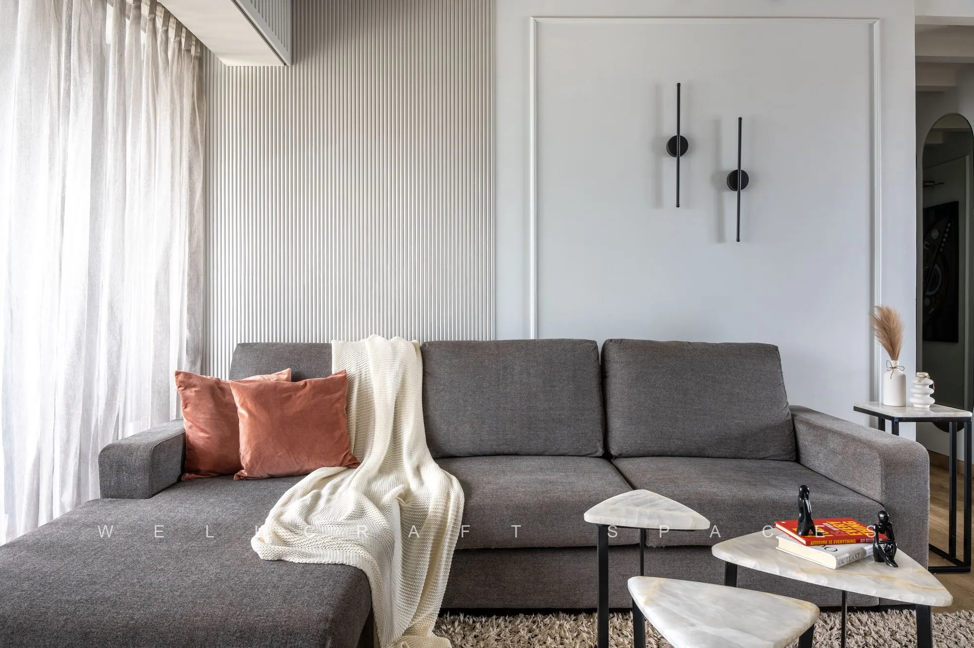 Grey L-shaped sectional with a cream throw and rust cushion, a round marble side table and sheer white curtains filtering daylight.