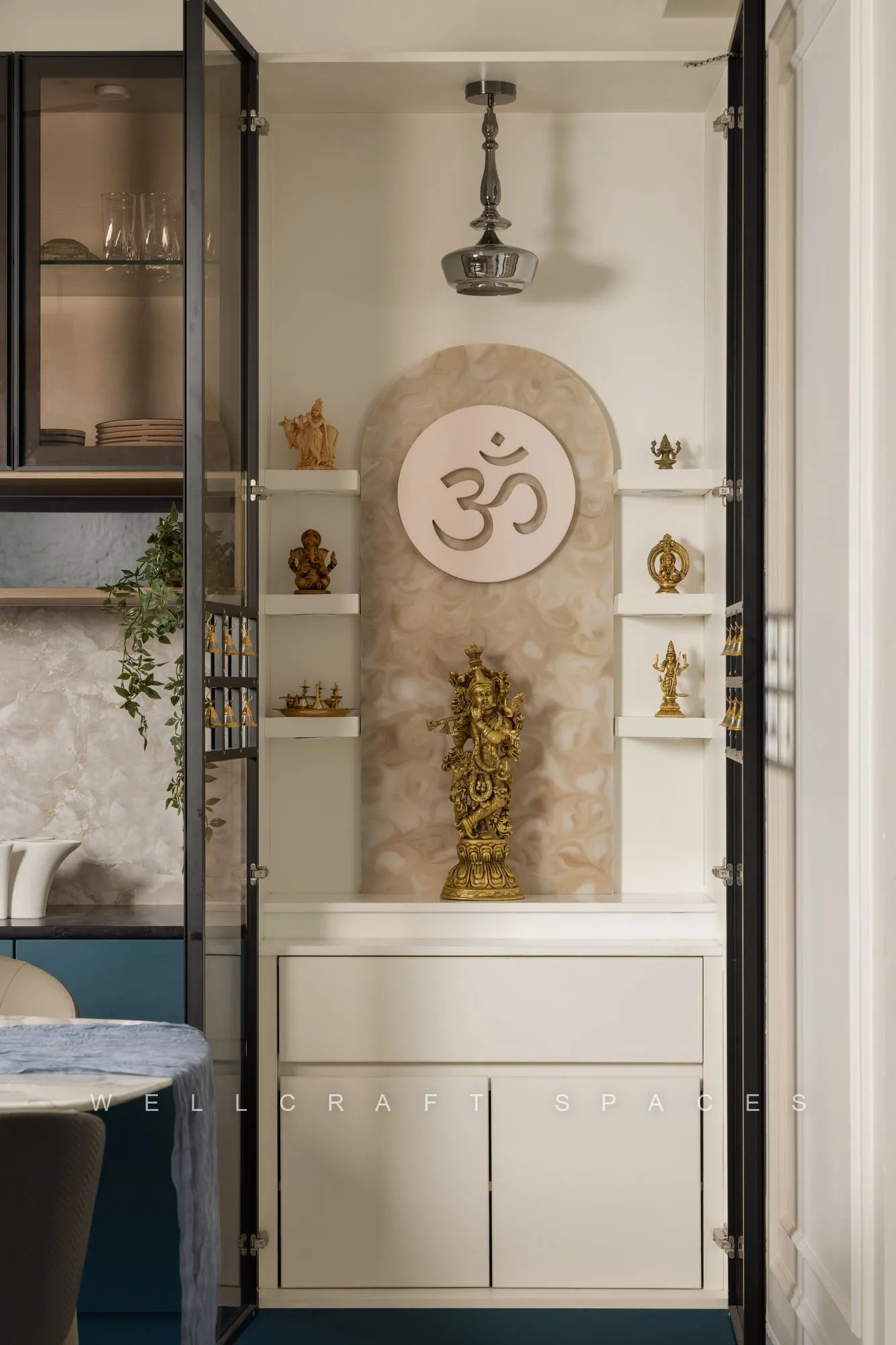 Pooja unit behind black-framed glass doors, with a carved Om backdrop, marble-toned platform and brass idols and lamps on the shelves.