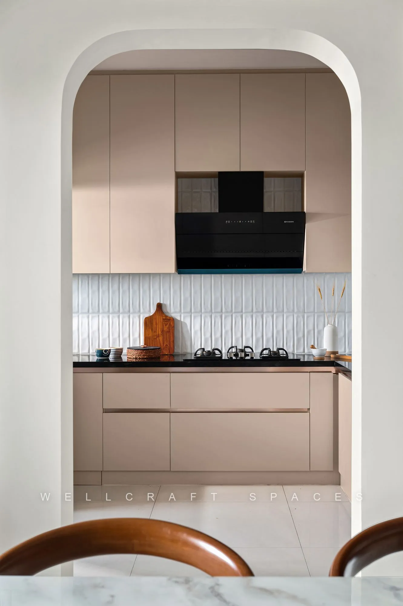 Kitchen glimpsed through a rounded arch, showing blush cabinetry, a black chimney and a fluted white tile backsplash.