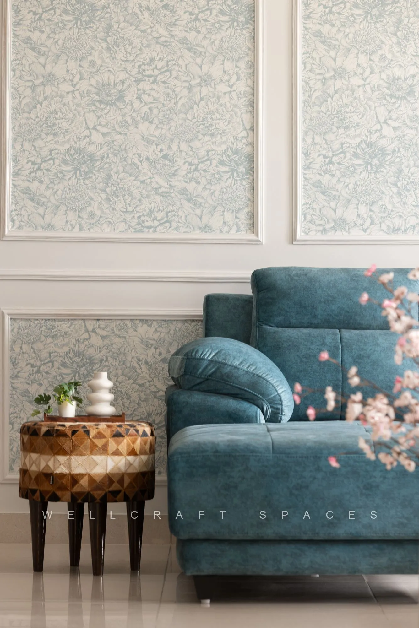 Close view of the teal sofa with a blossom-print cushion and a carved wood-and-brass side table against pale damask wall panels.