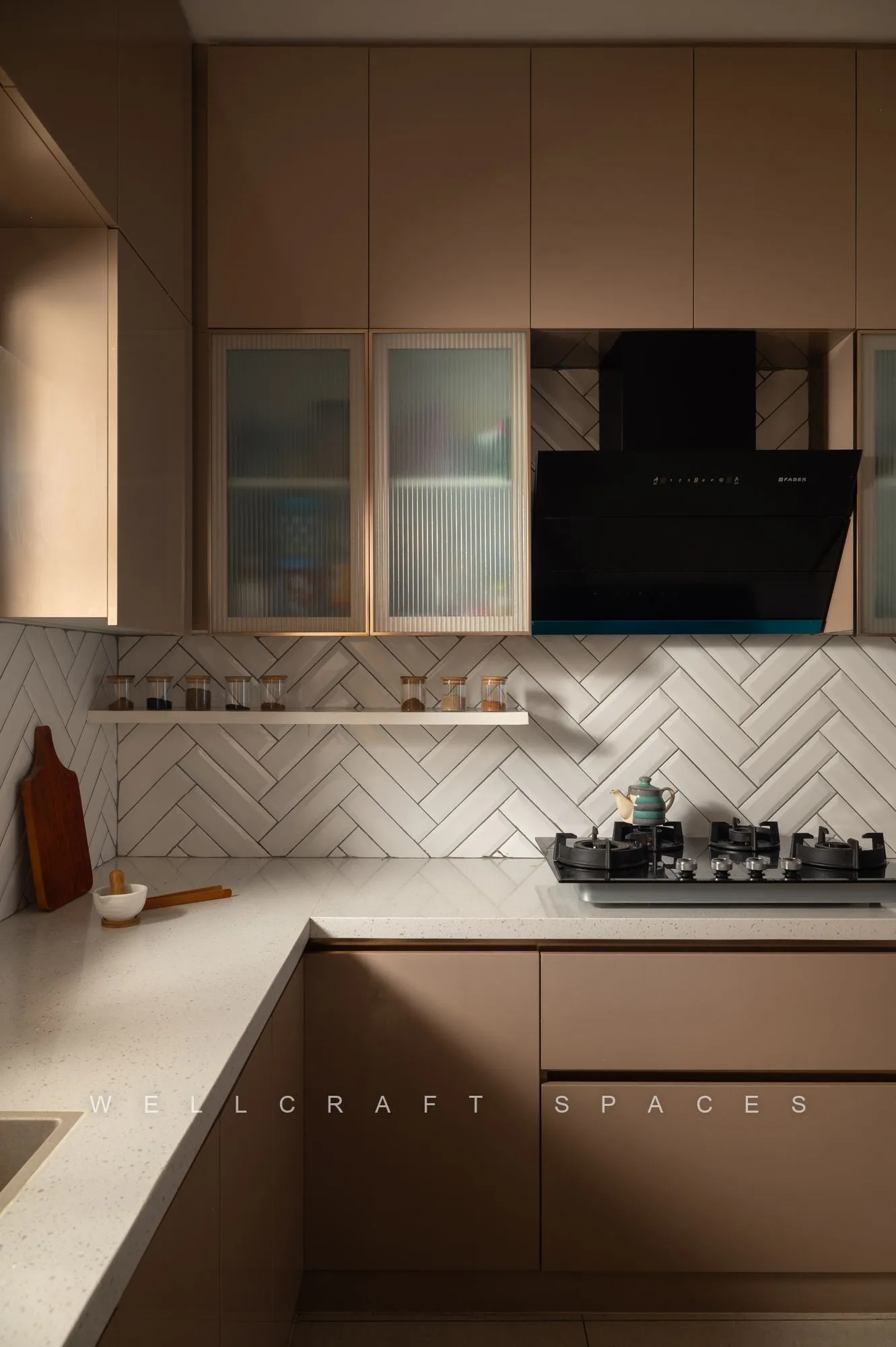L-shaped kitchen in matte blush-taupe cabinetry with a white herringbone tile backsplash, quartz counter and glass-fronted upper units.