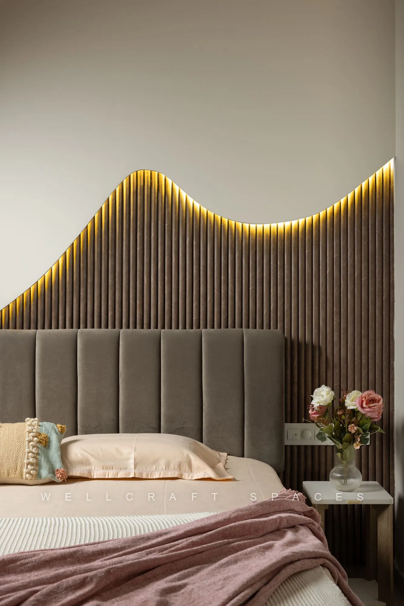 Master bed against a fluted taupe headboard wall with a wave-shaped backlit cove and a grey channel-tufted bedhead, dressed in blush linen.