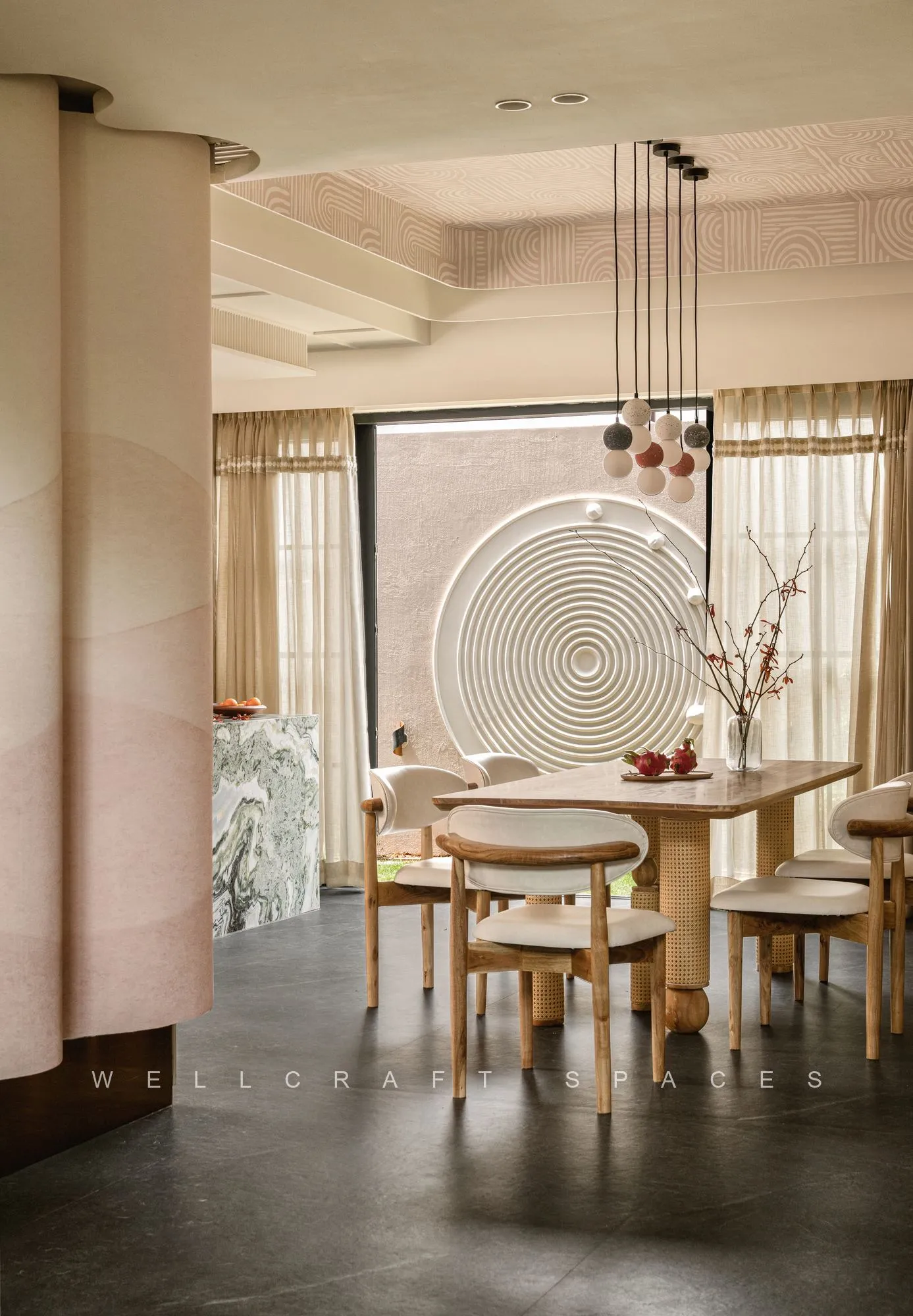 Dining area with a pale round table and timber chairs beneath a cluster of glass pendants, framed by a pink plaster arch and a decorative wall platter.