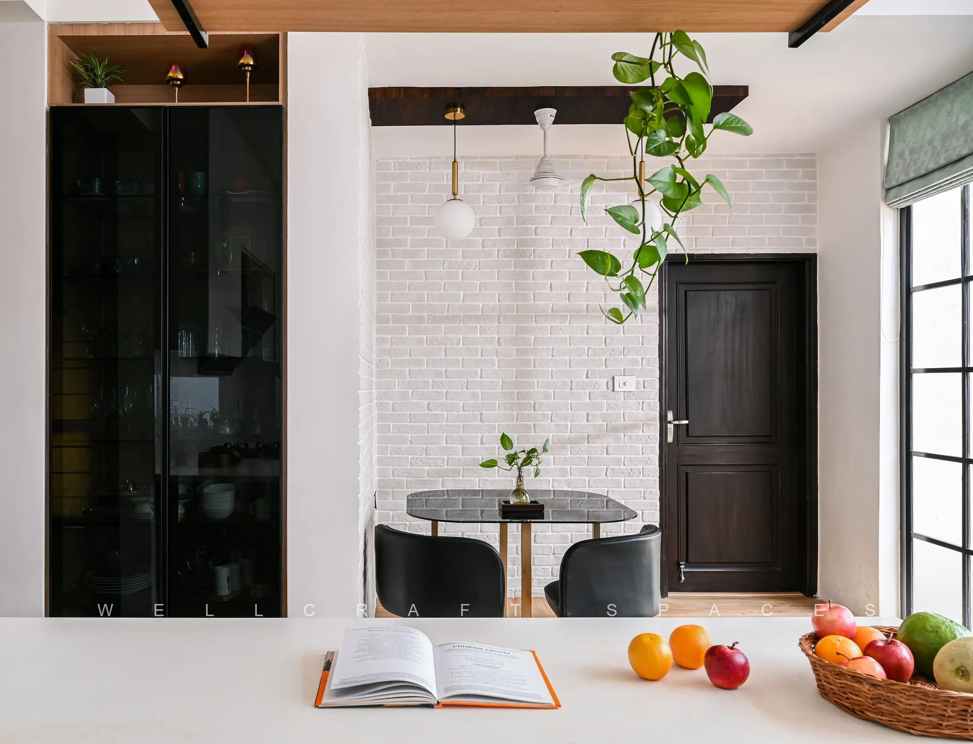 Dining corner against an exposed white brick wall, with a round marble-top table, black cane-back chairs and a glossy black sideboard under a slim pendant.