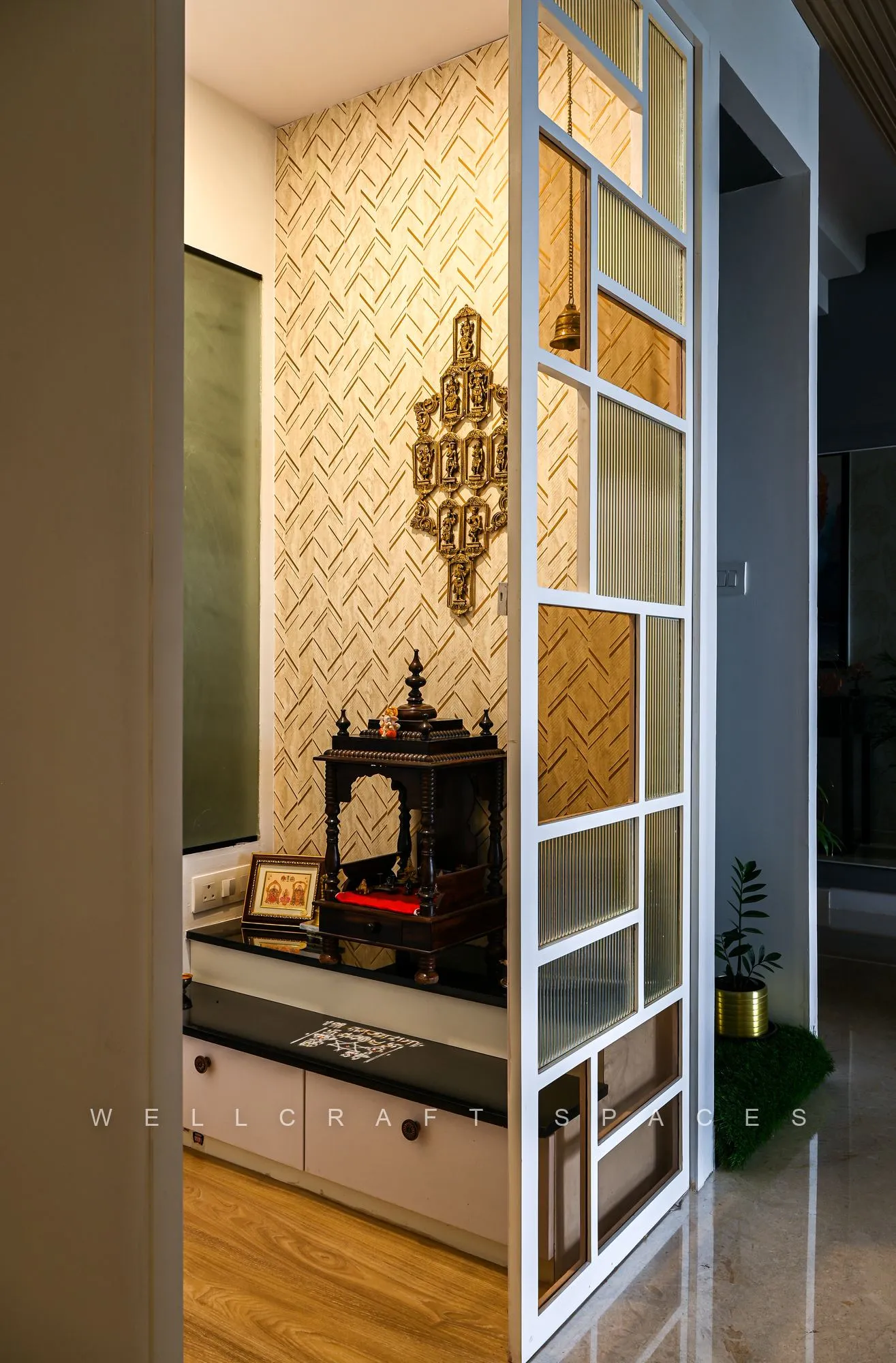 Pooja unit in a carved wooden mandir within a white grid frame, set against a gold patterned wall panel above warm timber flooring.