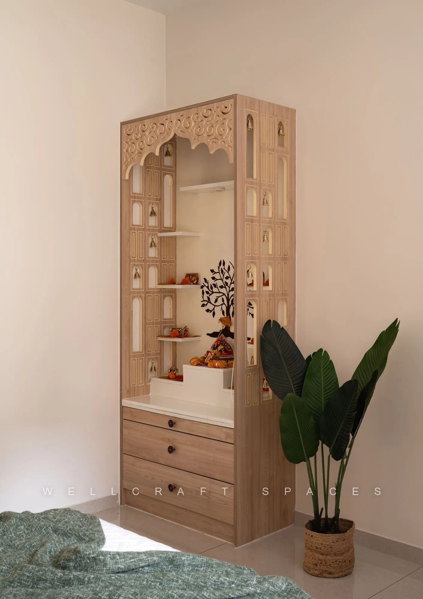 Freestanding pooja unit in carved light wood with a fretwork canopy, open shelves for idols and drawers below, beside a potted palm.