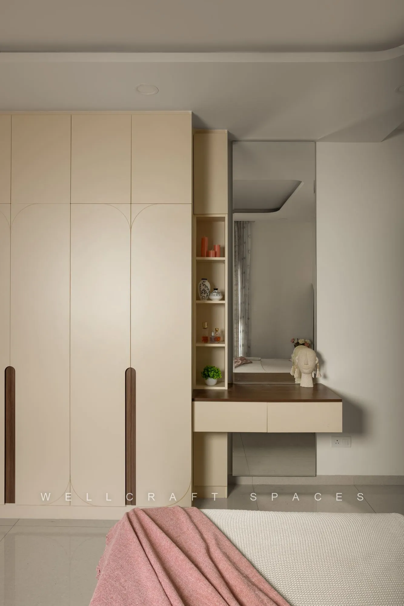 Ivory floor-to-ceiling wardrobe with slim walnut handles and an attached open dresser shelf, beside the blush-covered bed.