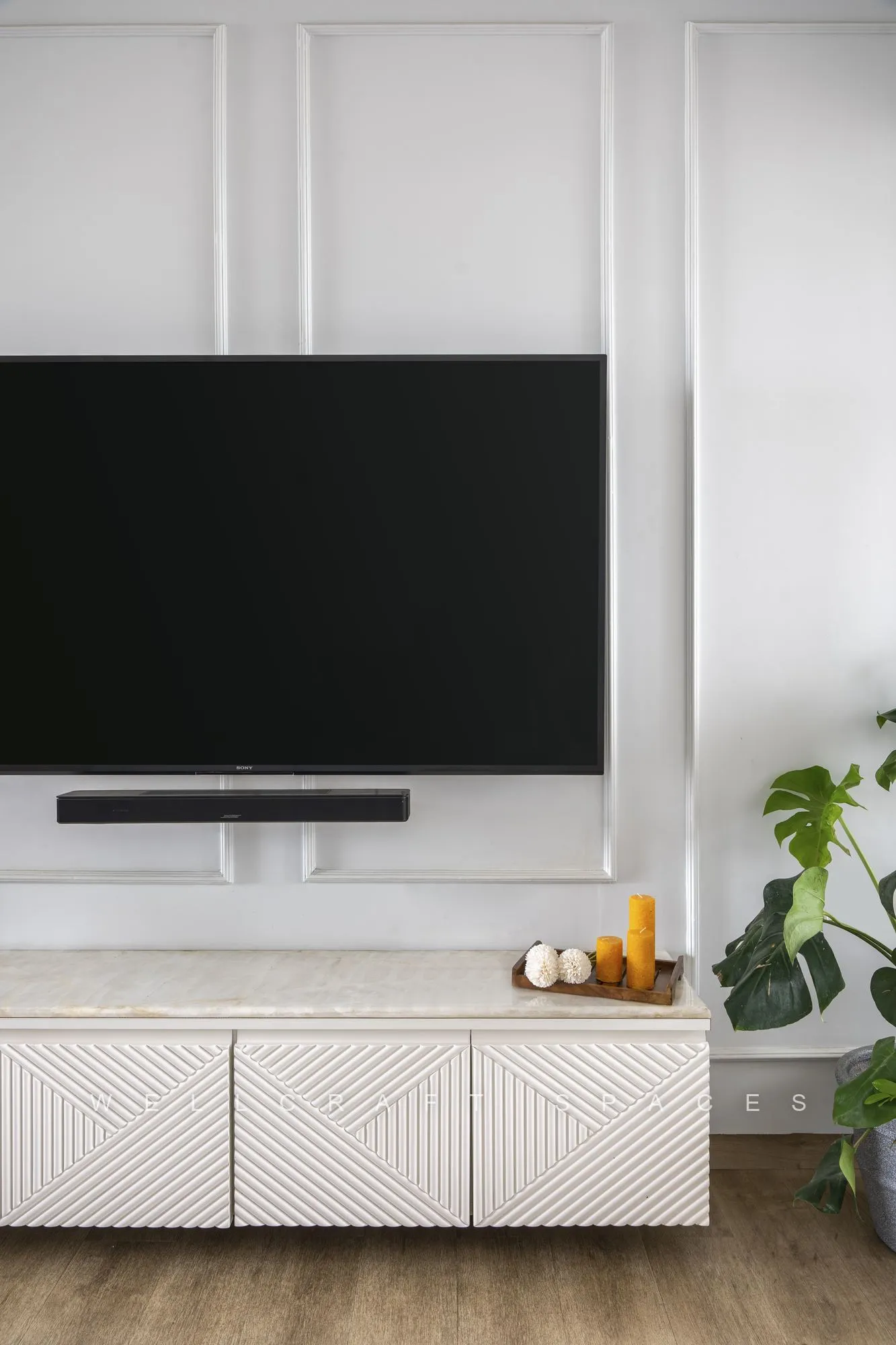 Television console detail in textured ivory with a soundbar, trailing greenery and small ceramic objects against the panelled wall.