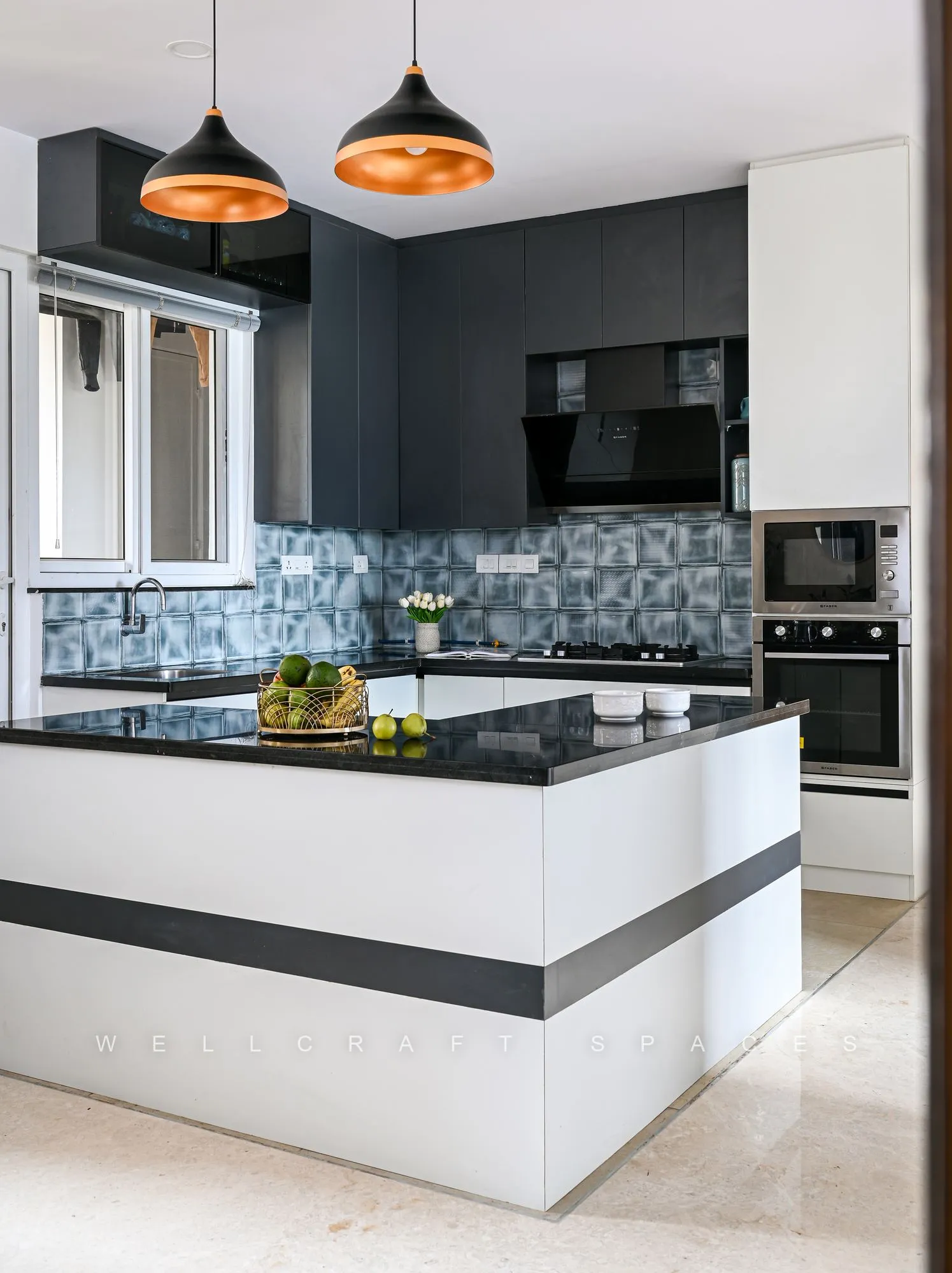 Kitchen island in white with a black granite top, copper dome pendants, patterned grey tile backsplash and built-in oven and microwave.