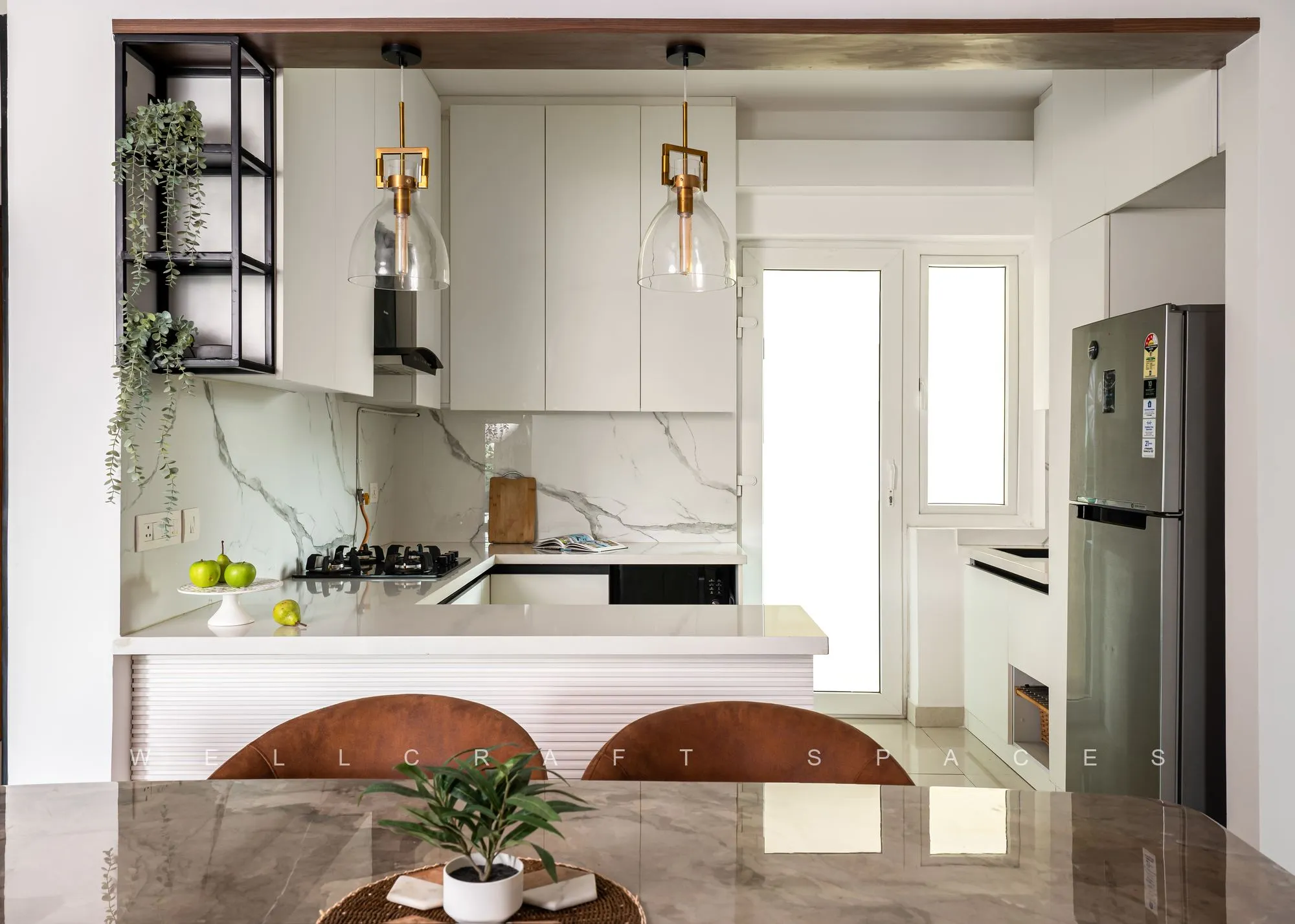 Kitchen seen across the dining table, with white shutters, a marble backsplash, black open shelves and brass pendant lights over the counter.
