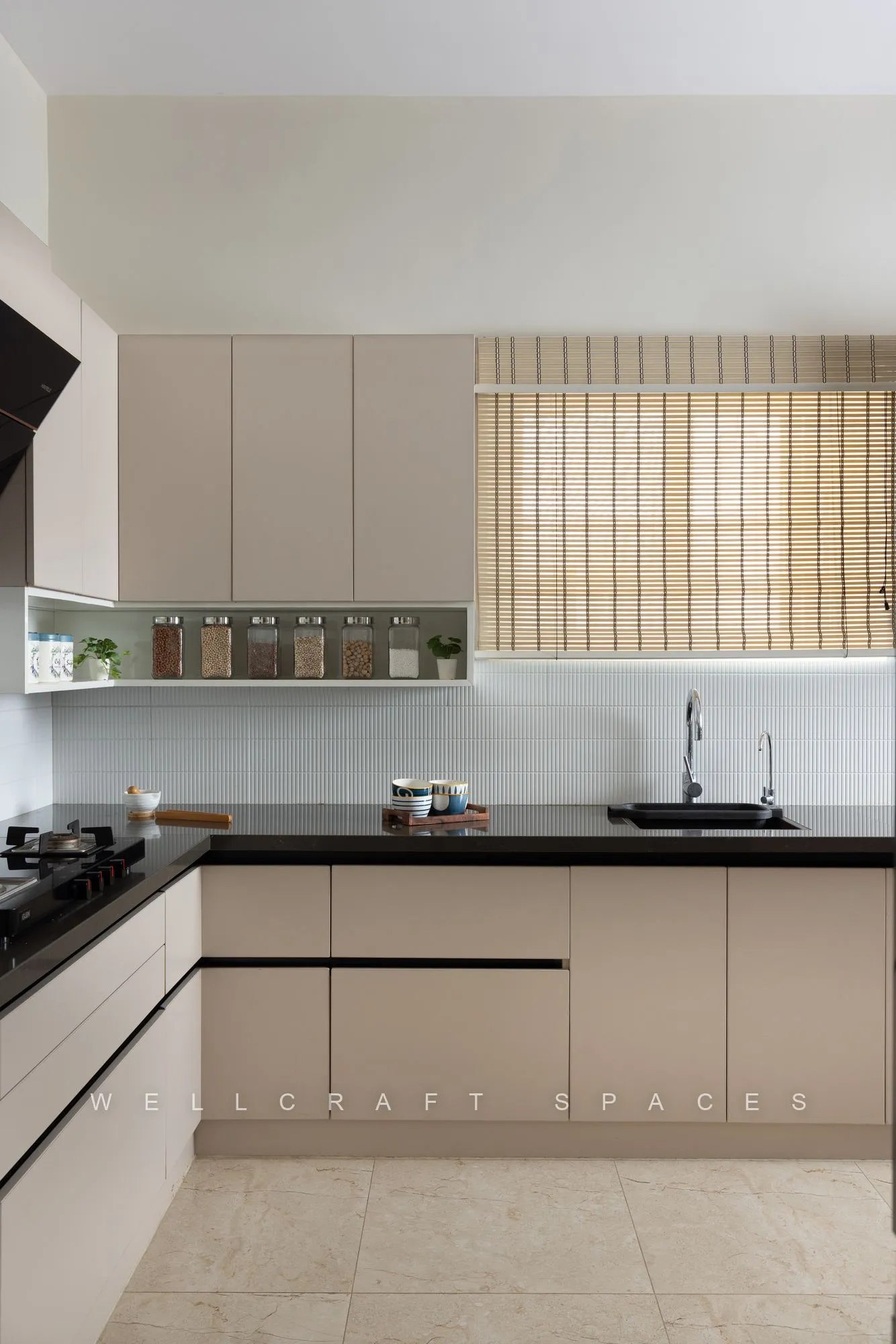 L-shaped kitchen in matt greige shutters with black granite counters, a white tiled backsplash, bamboo blinds and a built-in hob and sink.
