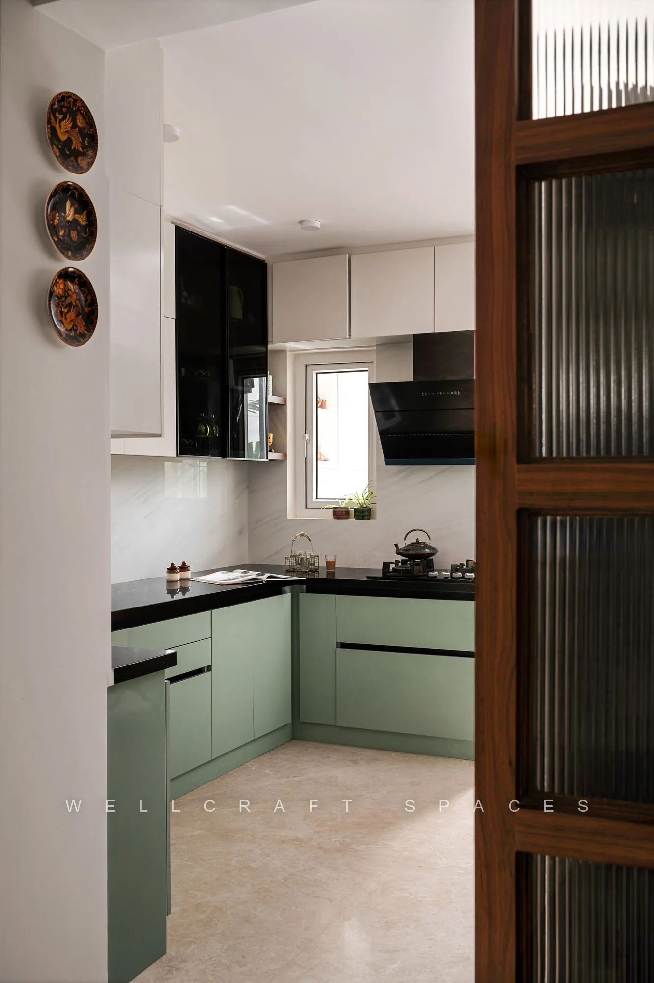 Sage green kitchen viewed past a fluted glass and walnut door, with black counters, a built-in hob and decorative plates on the wall.