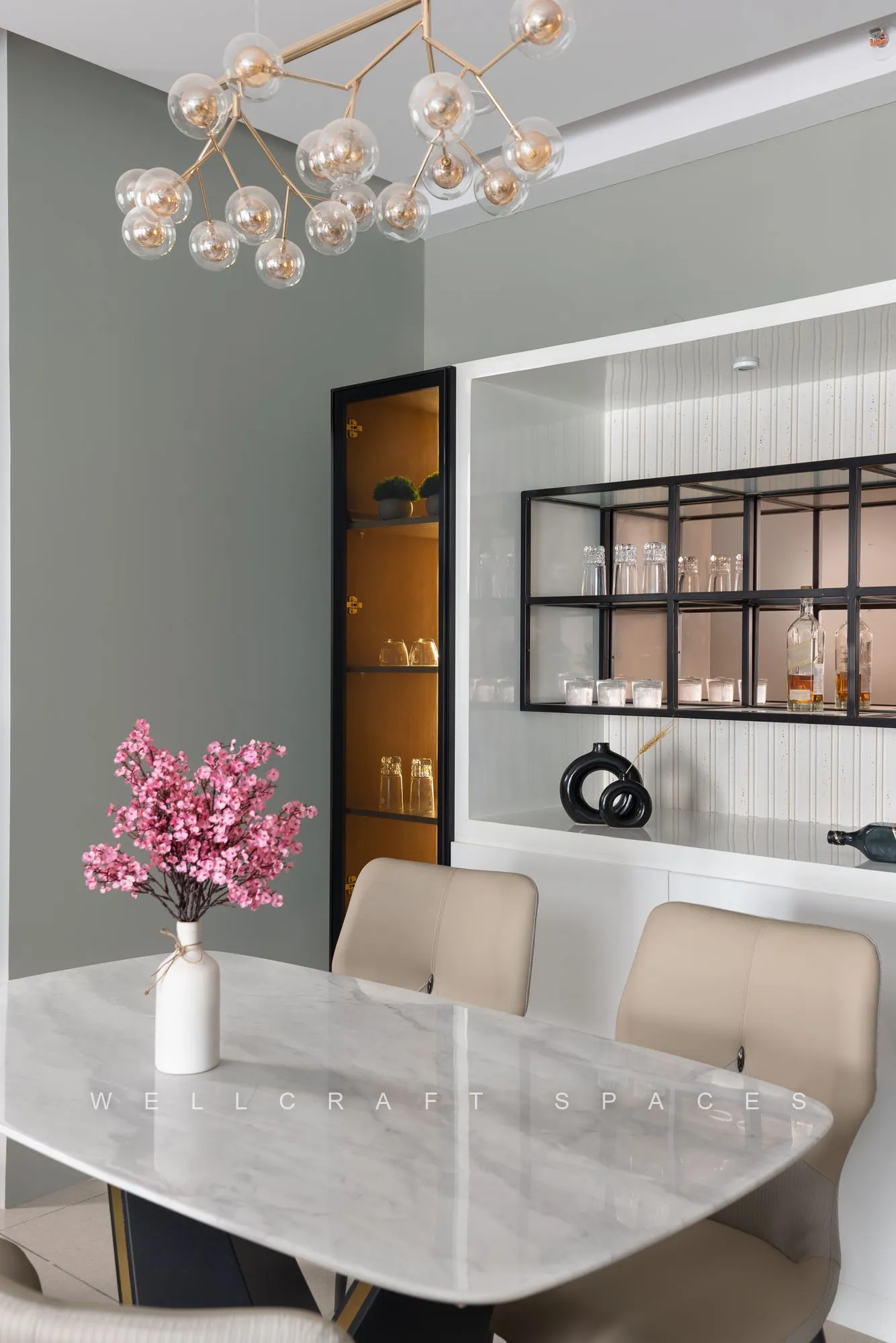 Marble-topped dining table with cream upholstered chairs beneath a brass sputnik chandelier, next to a black-framed glass crockery unit.