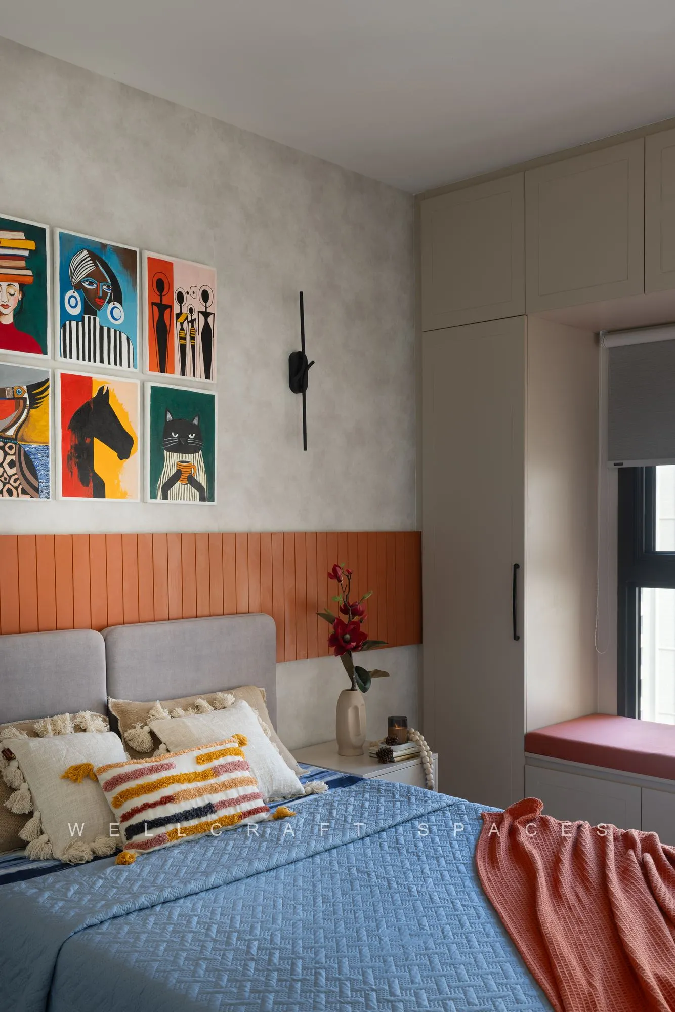 Master bed with a rust upholstered headboard band, blue quilt and striped cushions, under a gallery wall of colourful framed prints.