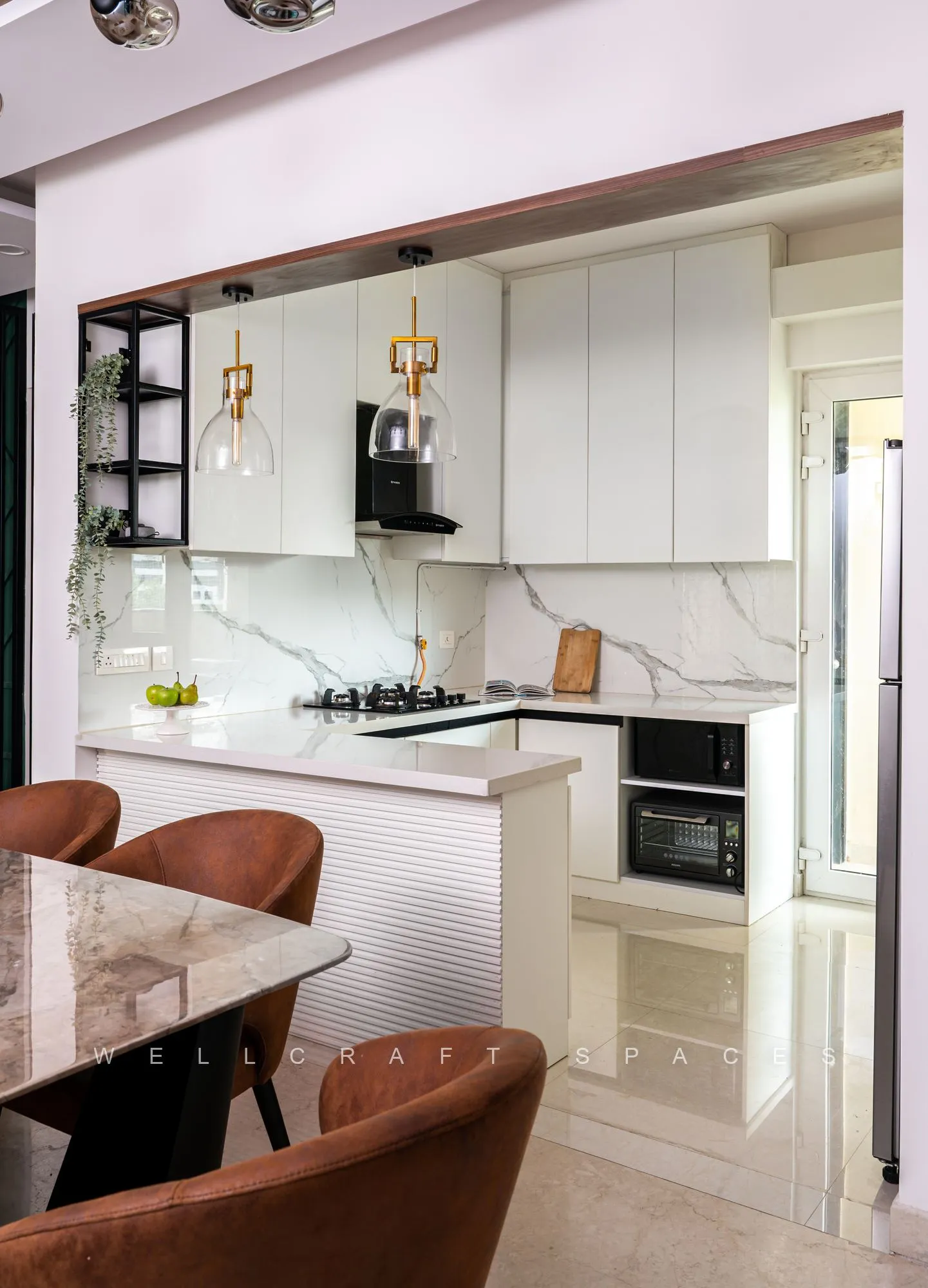 White kitchen with marble-veined counters and backsplash, a built-in hob and oven, brass-and-glass pendants and rust leather chairs in the foreground.
