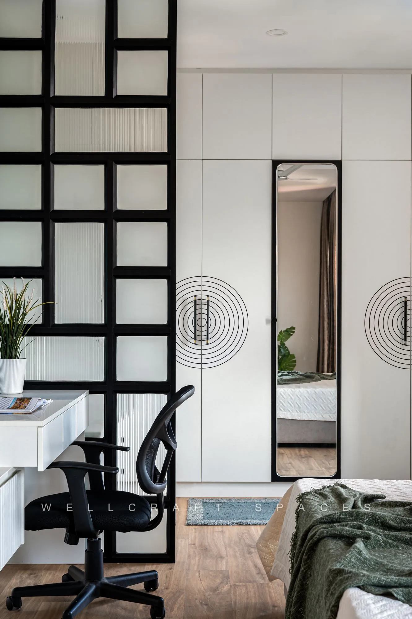 Bedroom divided by a black grid glass partition, with a white work desk, mesh chair and a full-length mirror on the wardrobe front.