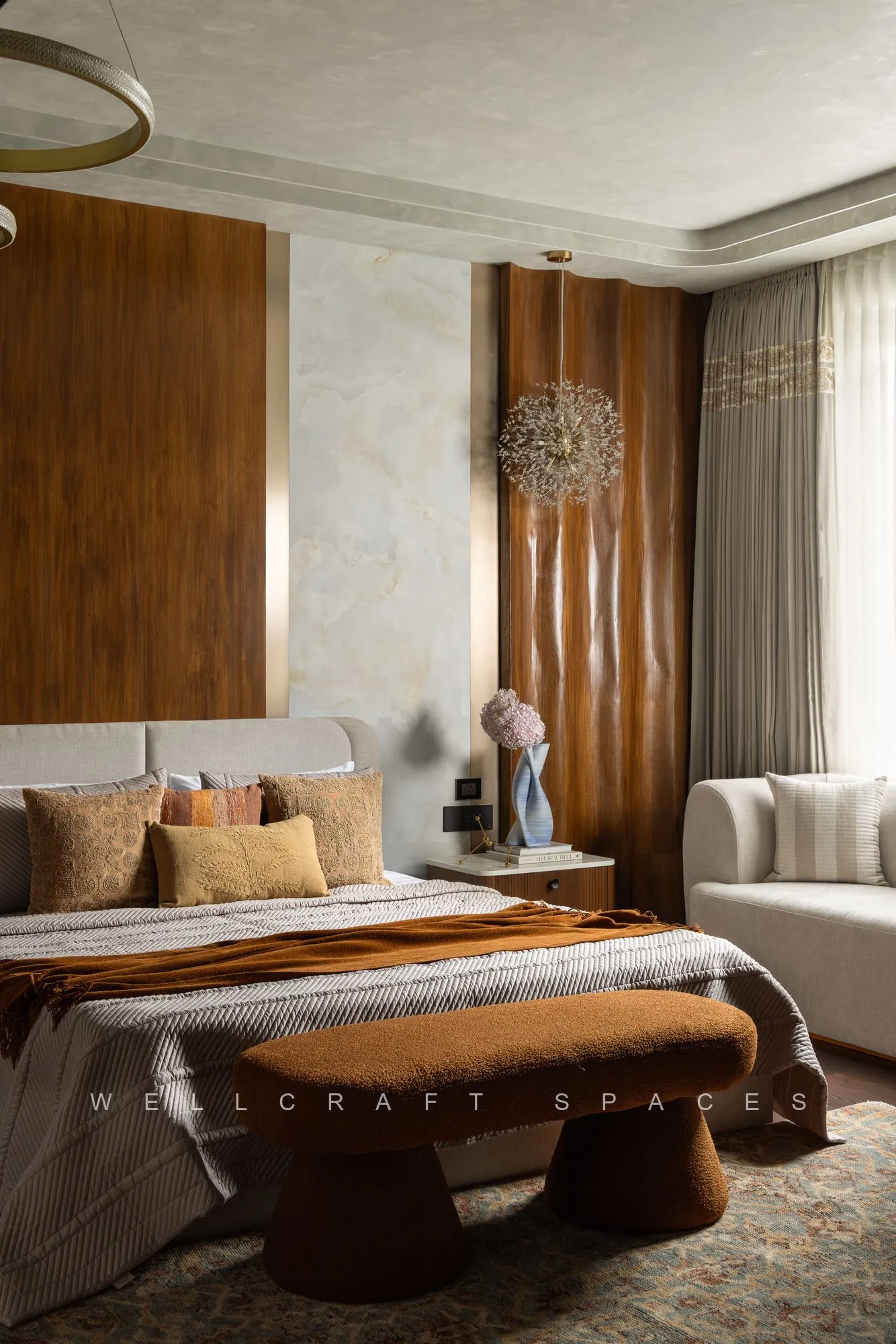 Bedroom with fluted walnut panelling and floor-length drapes, a cream upholstered headboard, striped throw and a sculptural curved bench.