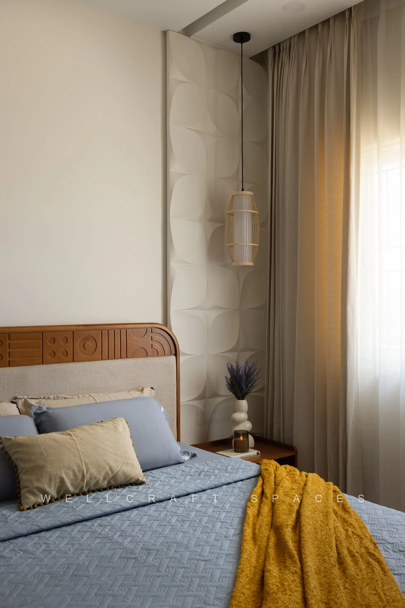 Master bed with a quilted cream headboard, blue quilt and mustard throw, lit by a woven pendant beside the curtained window.