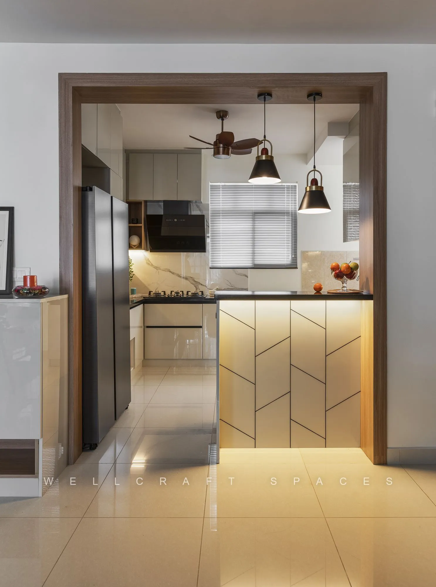 Warmly lit kitchen framed by a teak-clad opening, showing herringbone-panelled cabinet fronts and pendant lights over the counter.