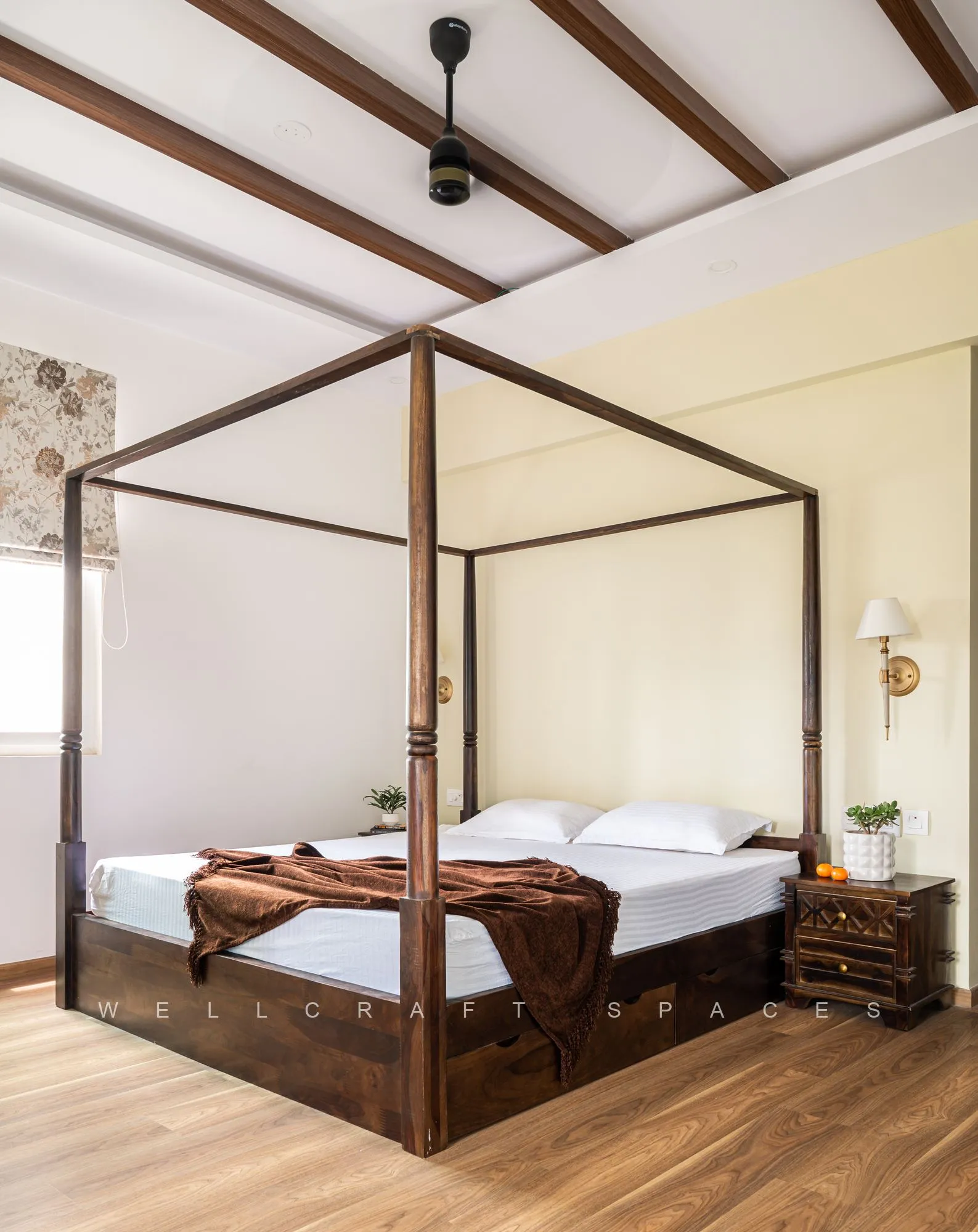 Master bedroom with a dark teak four-poster bed under exposed timber beams, a chocolate throw, a brass wall sconce and a floral-patterned roman blind.