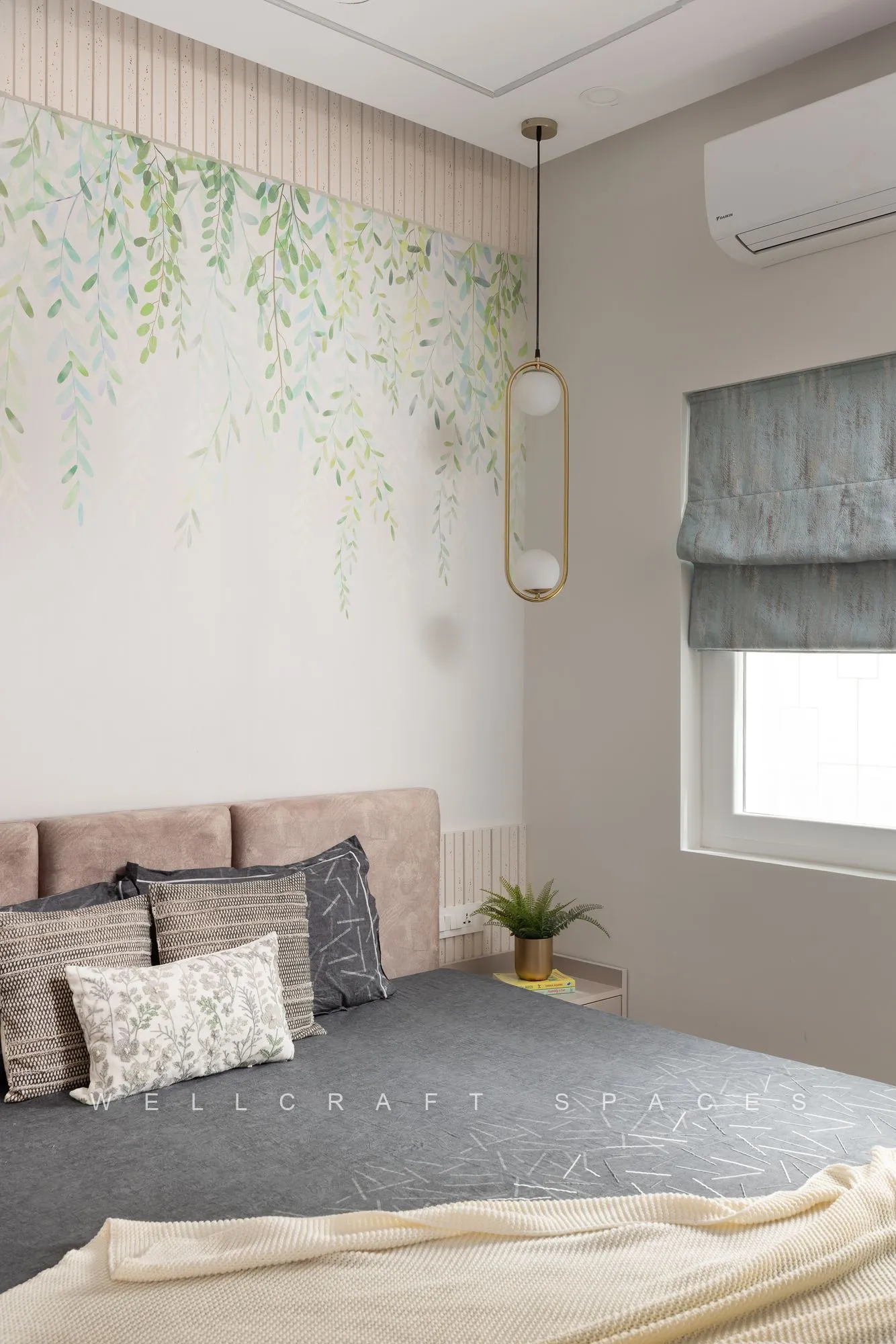 Bedroom with a blush fabric headboard, grey bedspread, trailing-foliage wallpaper and a slim gold pendant beside a fabric roman blind.