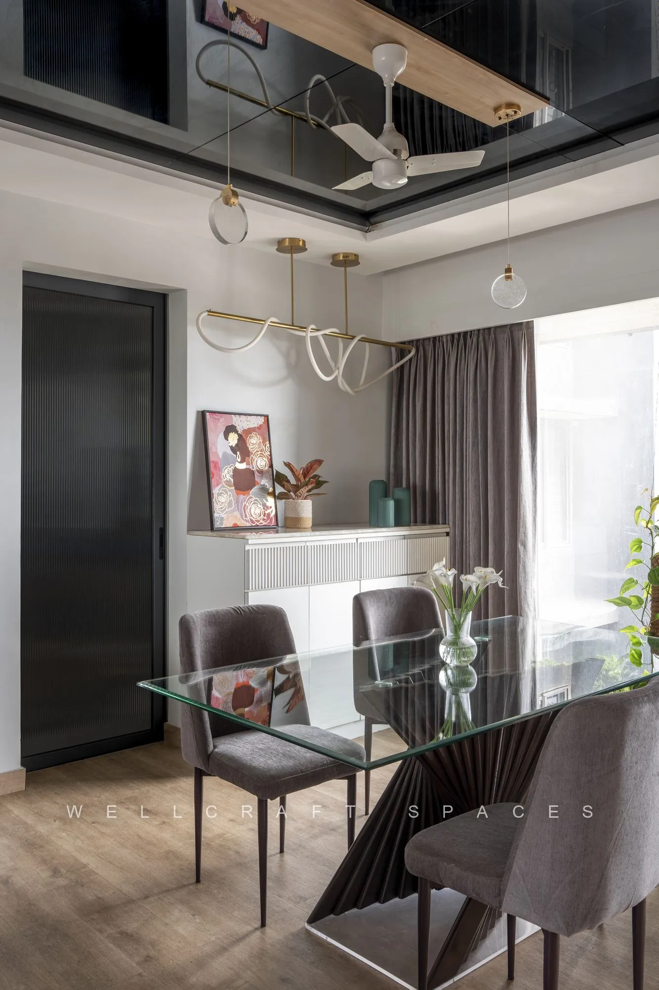 Glass-top dining table with grey chairs beneath a sculptural brass ring pendant, backed by a white fluted sideboard and a mirrored ceiling inset.