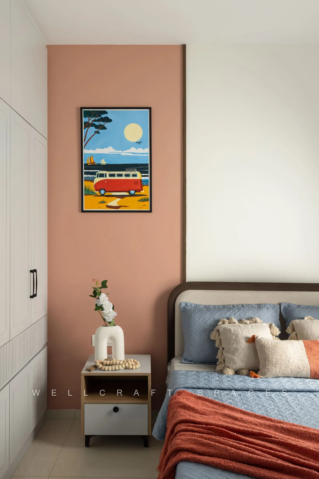 Bed with a teal headboard and coral throw beside a terracotta-painted wall hung with a bright seaside print.