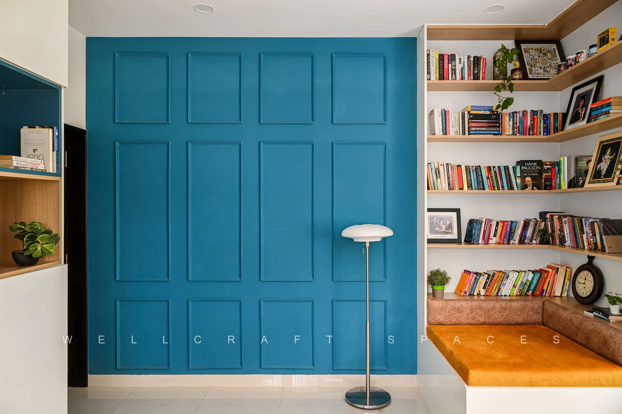 Library corner with a deep teal panelled wall, floor-to-ceiling walnut bookshelves, a cantilevered timber window seat and an arc floor lamp.