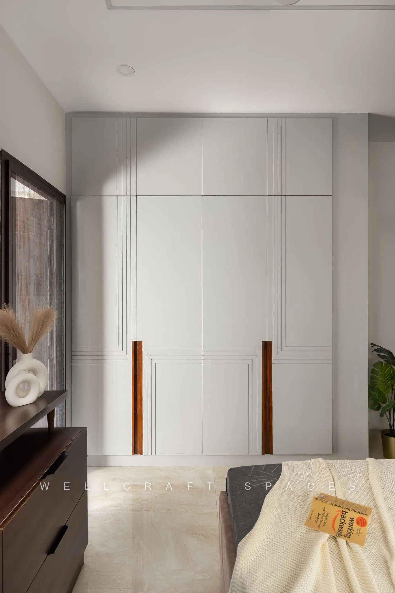 Wardrobe wall in ivory with slim tan leather pulls, beside a dark timber chest and a bed corner in soft neutral bedding.
