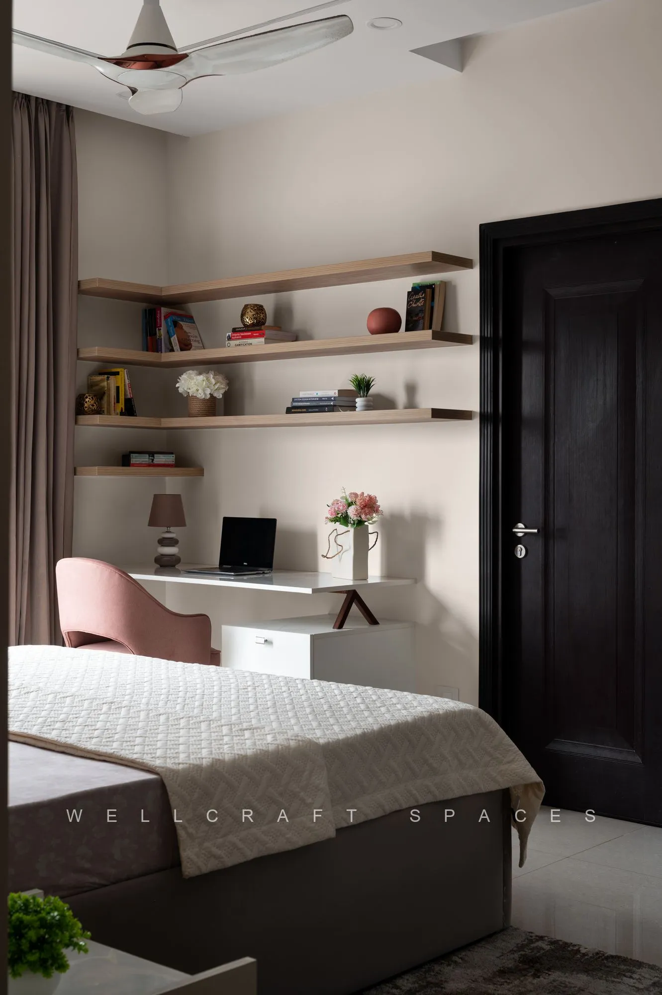 Compact study nook with floating wood shelves, a white desk, blush upholstered chair and a dark panelled door beside the bed.