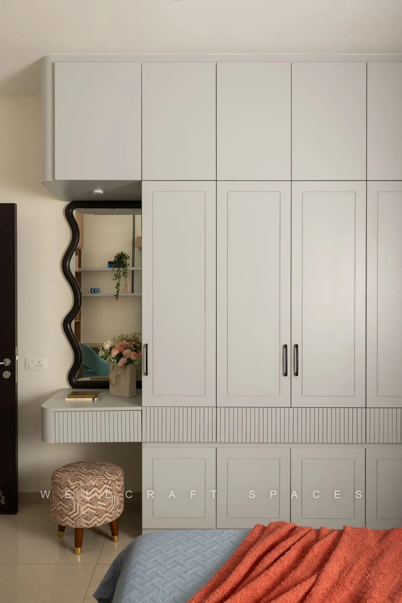Fluted cream wardrobe wall with a compact dressing ledge, arched mirror and a small upholstered stool.