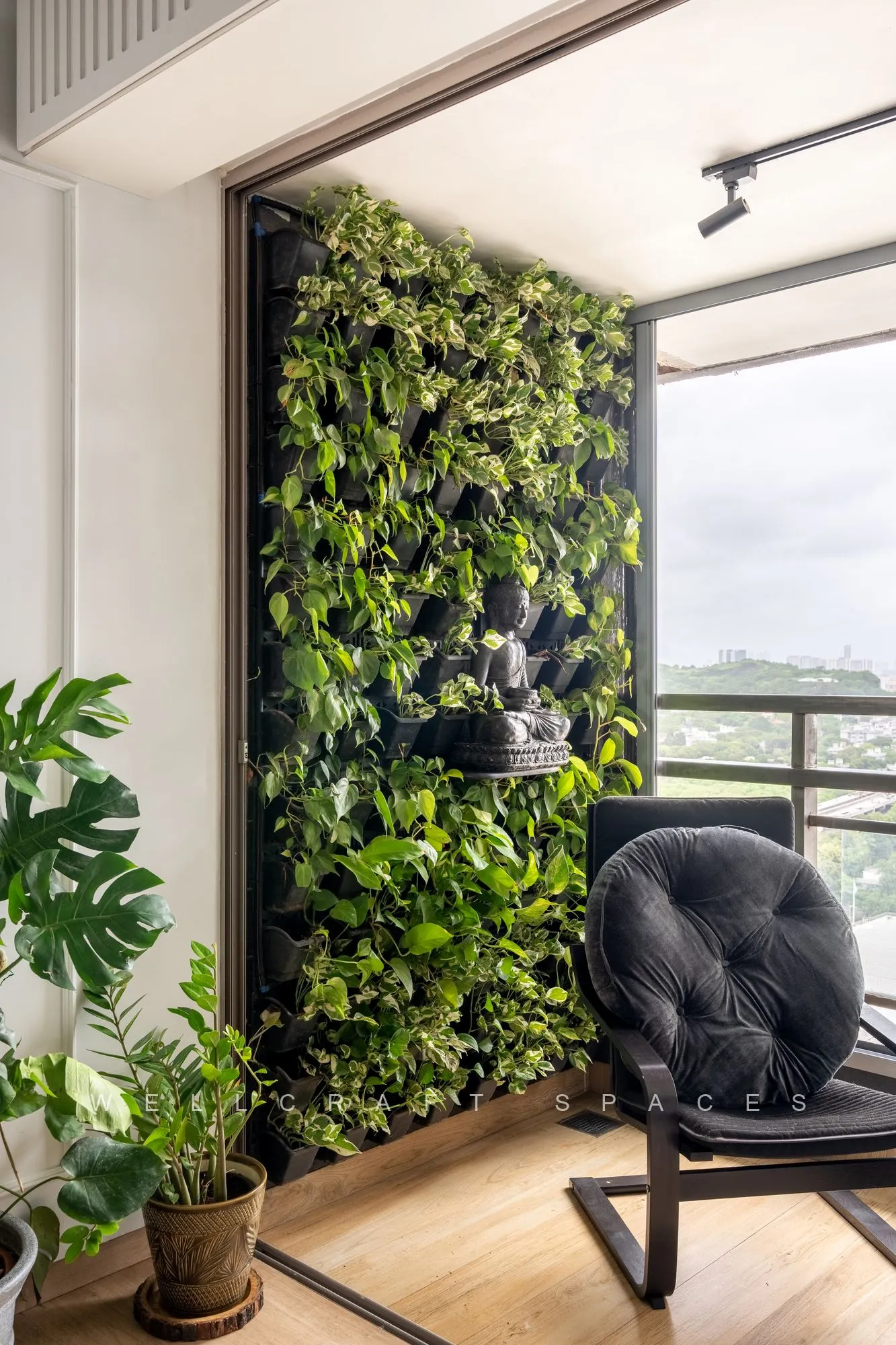 Enclosed balcony with a full-height artificial green wall, a black round lounge chair and clustered potted plants beside the glazing.