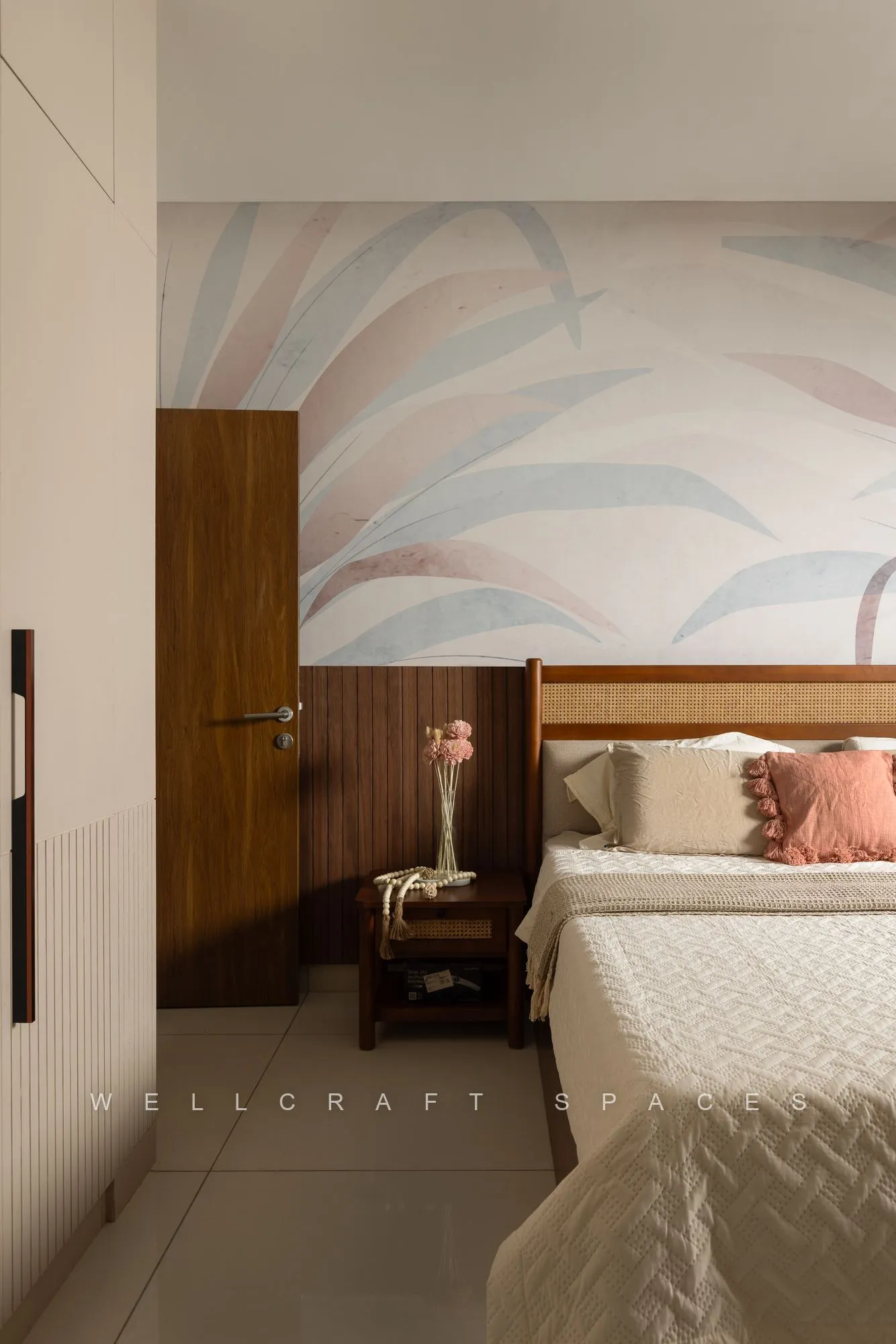 Bed with a teak headboard against a pastel watercolour mural wall, with a wooden nightstand and a tall wardrobe by the door.