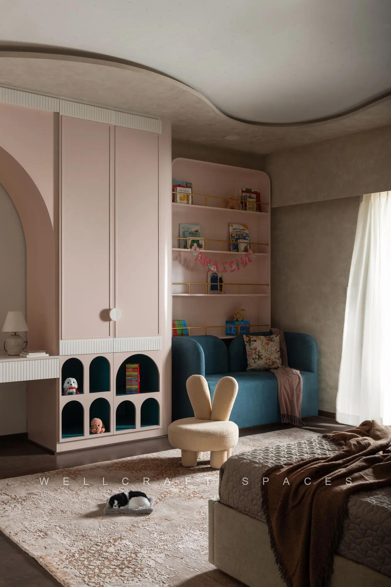 Kids room with a blush arched display unit, navy cushioned reading nook, open shelving and a soft patterned rug.