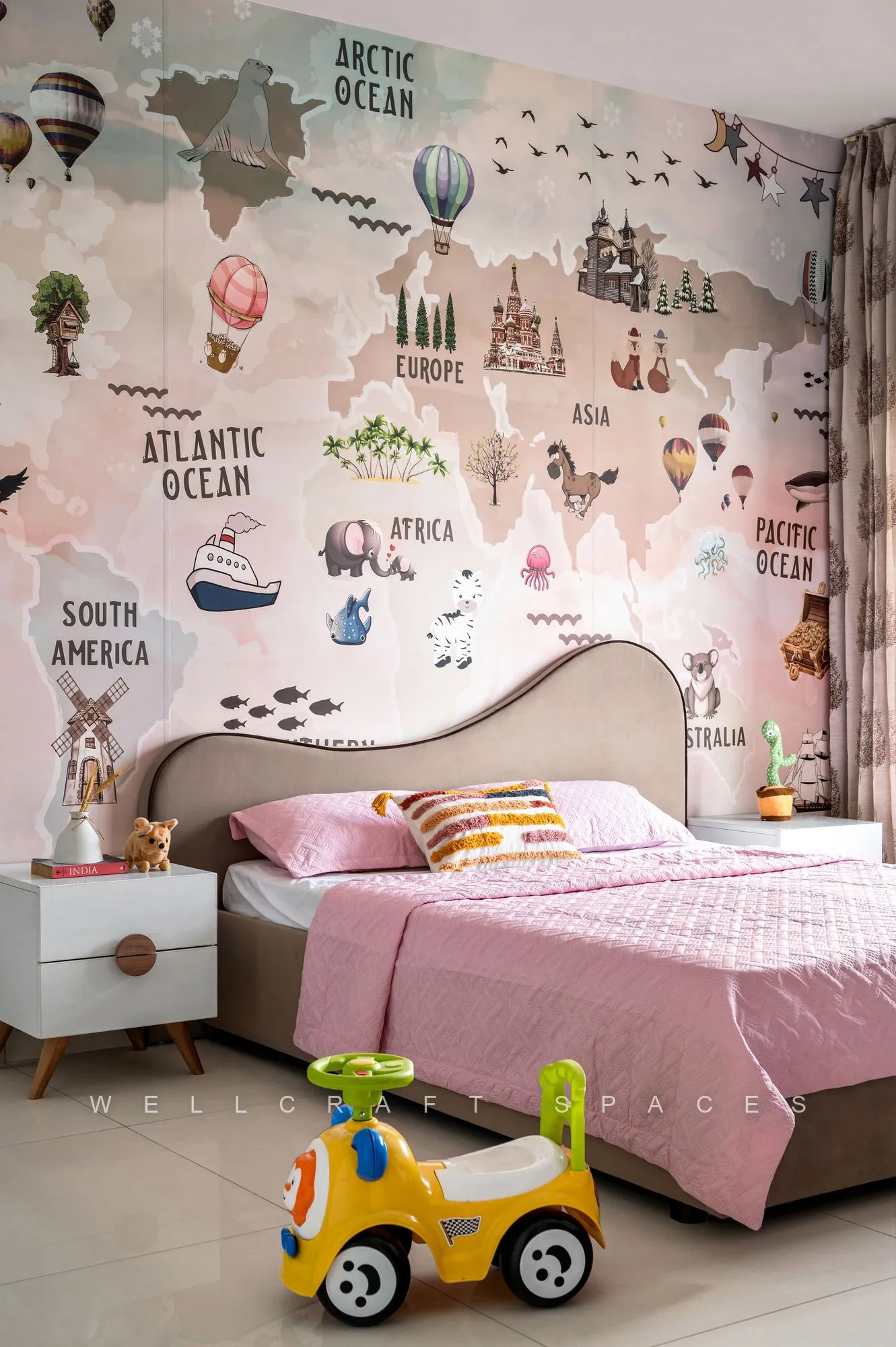 Child's bed with pink bedding and a curved headboard against a full-wall illustrated world-map mural, with a ride-on toy on the floor.