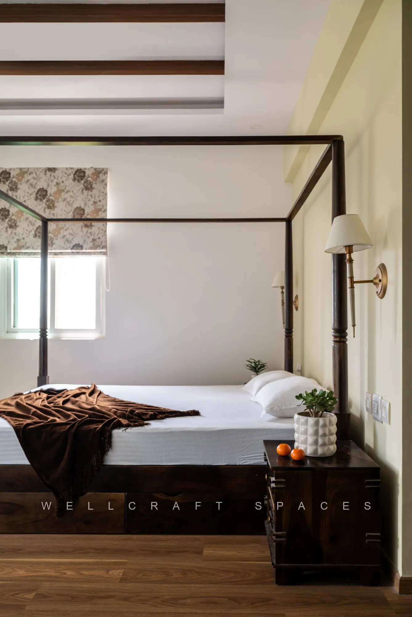 Four-poster bed detail with crisp white linen and a brown knitted throw, a black glossy bedside chest and a brass swing-arm lamp on the ivory wall.