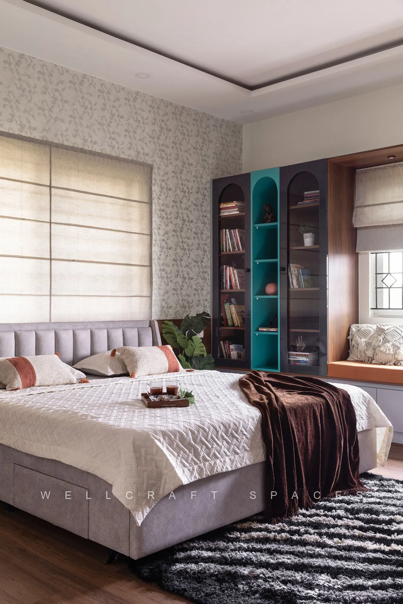 Master bedroom with a grey channel-tufted bed, quilted bedding and a chocolate throw, a teal open bookshelf unit and delicate patterned wallpaper behind.
