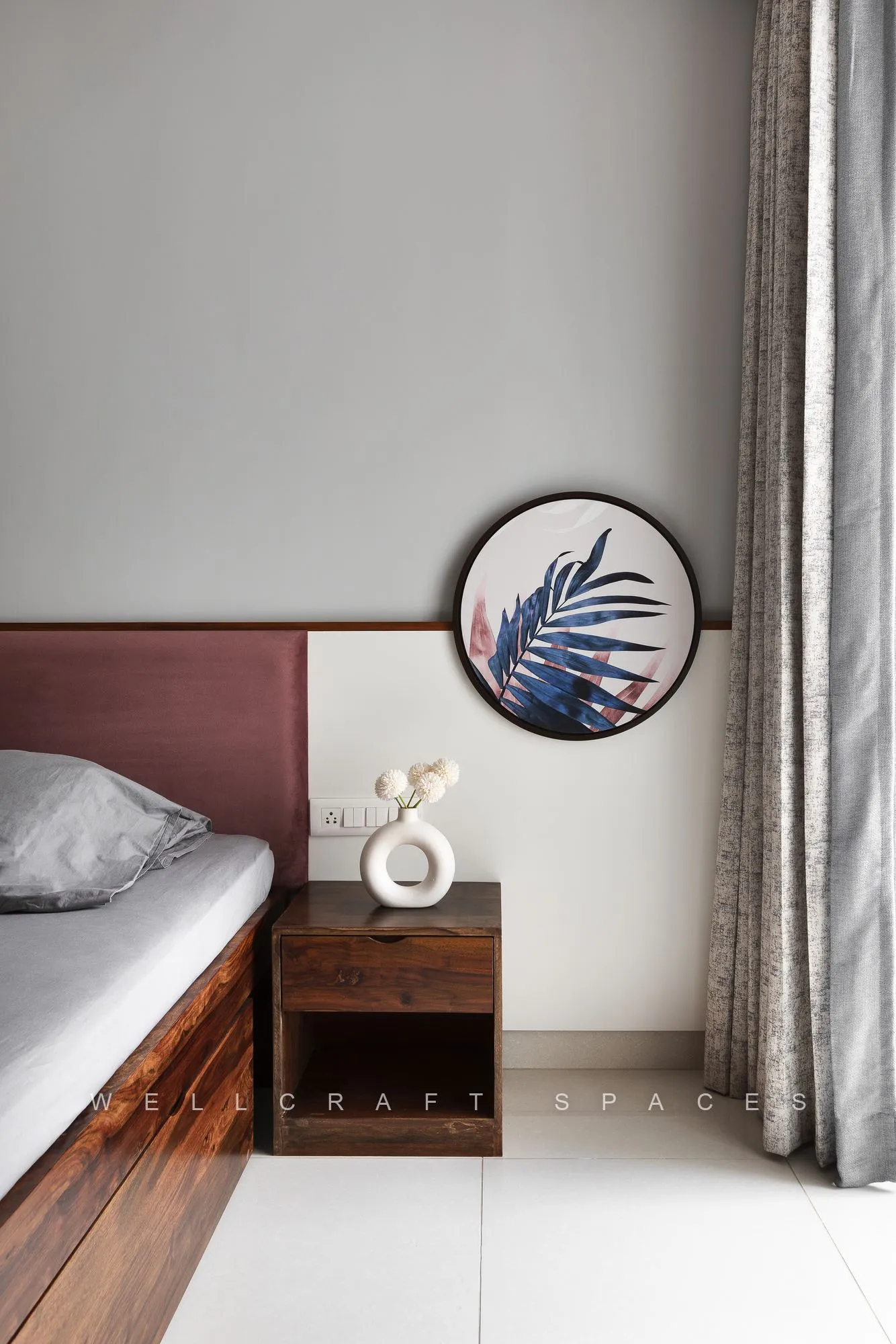 Bedside corner with a teak nightstand, a round framed palm-leaf print and the maroon headboard against a soft grey wall.