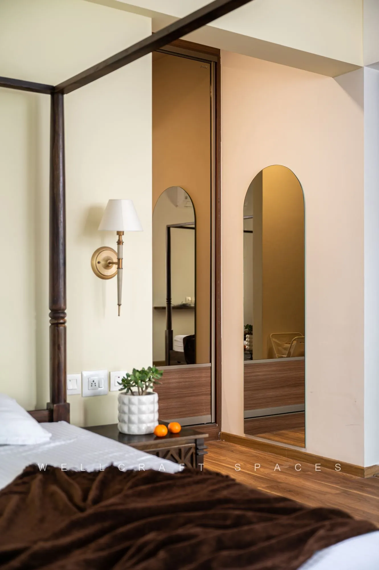 Bedroom with a full-height mirrored wardrobe reflecting an arched doorway, a brass wall sconce and a bed dressed in white with a brown throw.