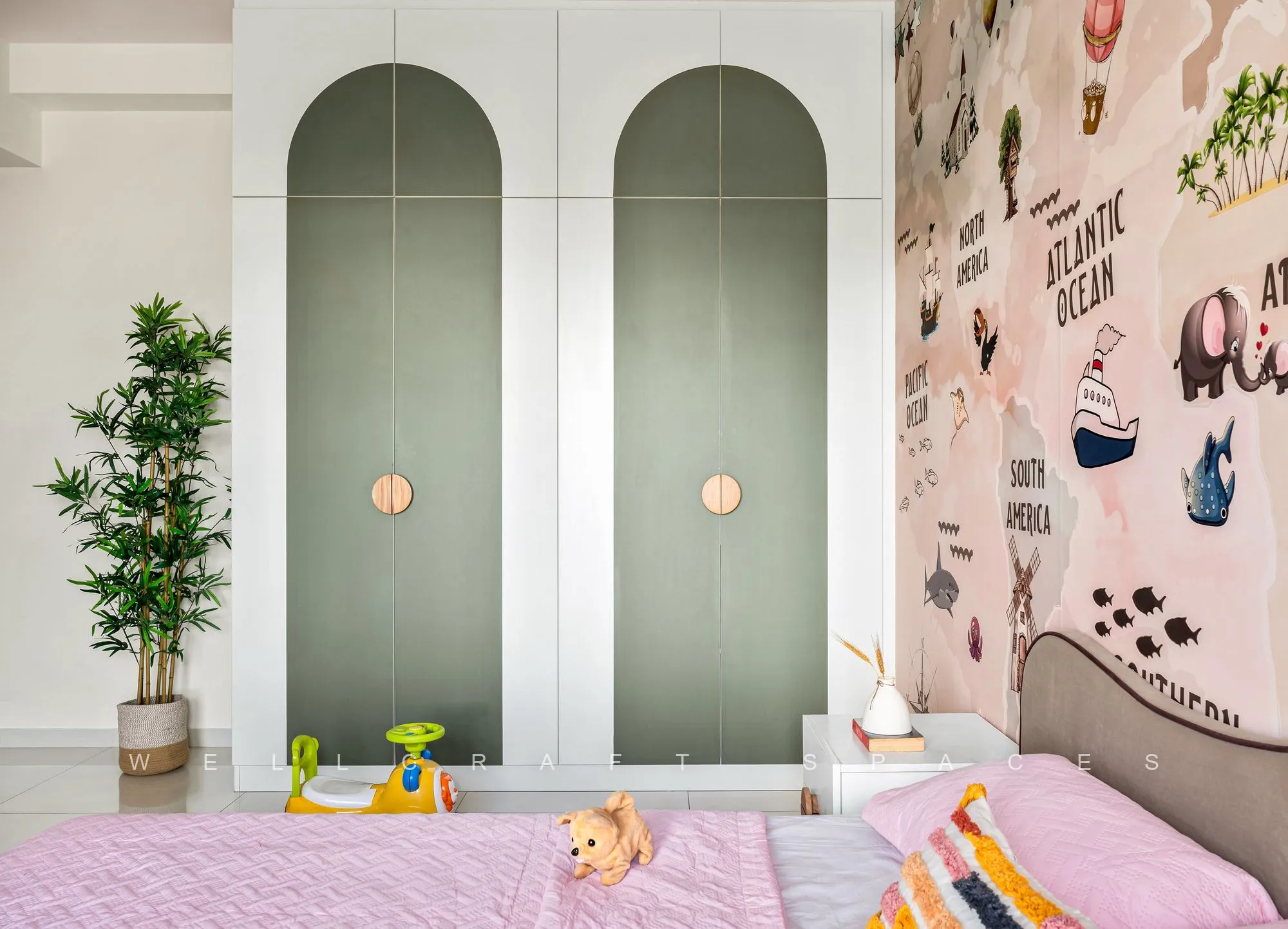 Sage-green arched wardrobe doors with brass pulls beside the world-map mural wall and a pink-covered bed.