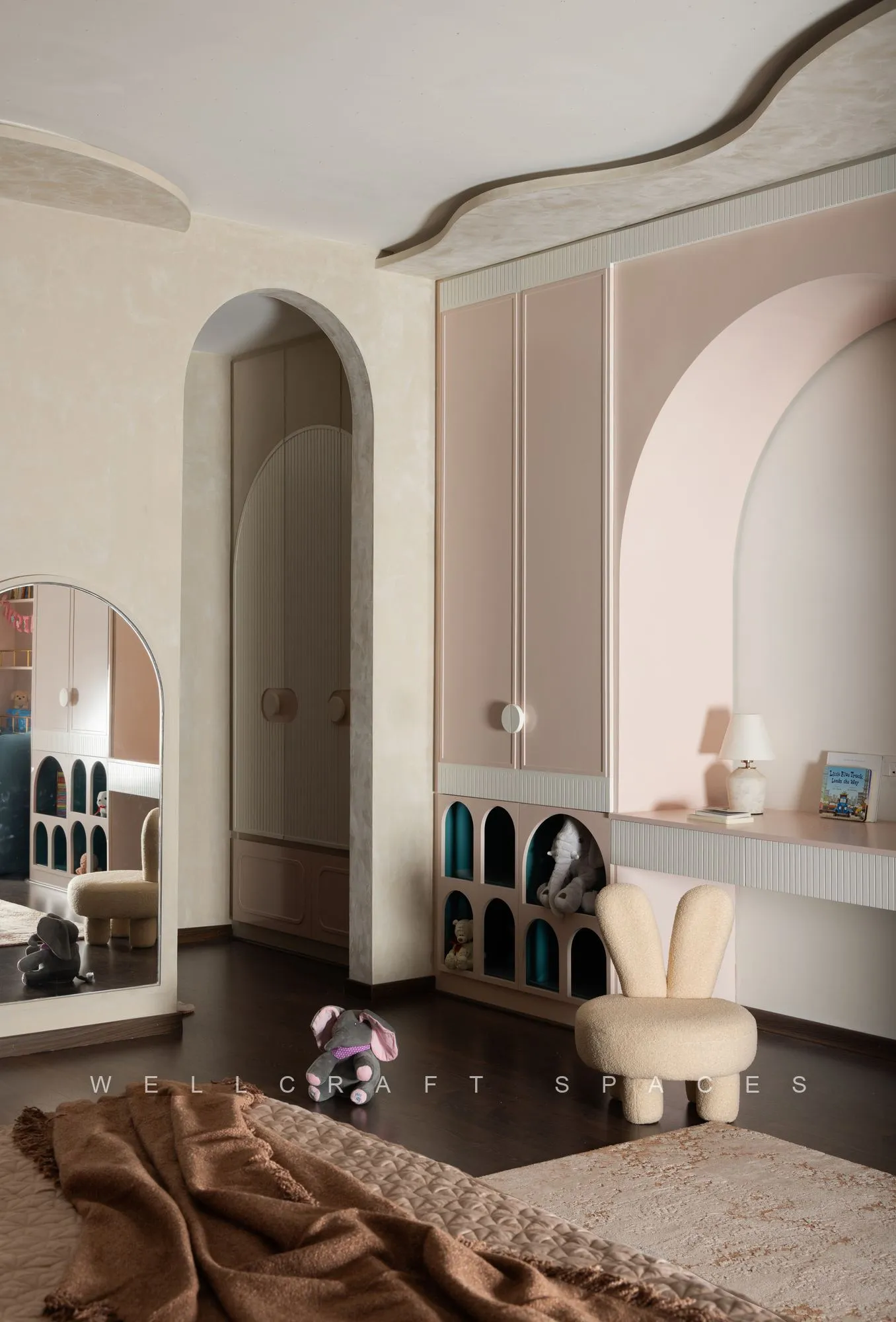 Kids room dressing corner with a pale arched vanity niche, round mirror, cream stool and a curved white wardrobe run.