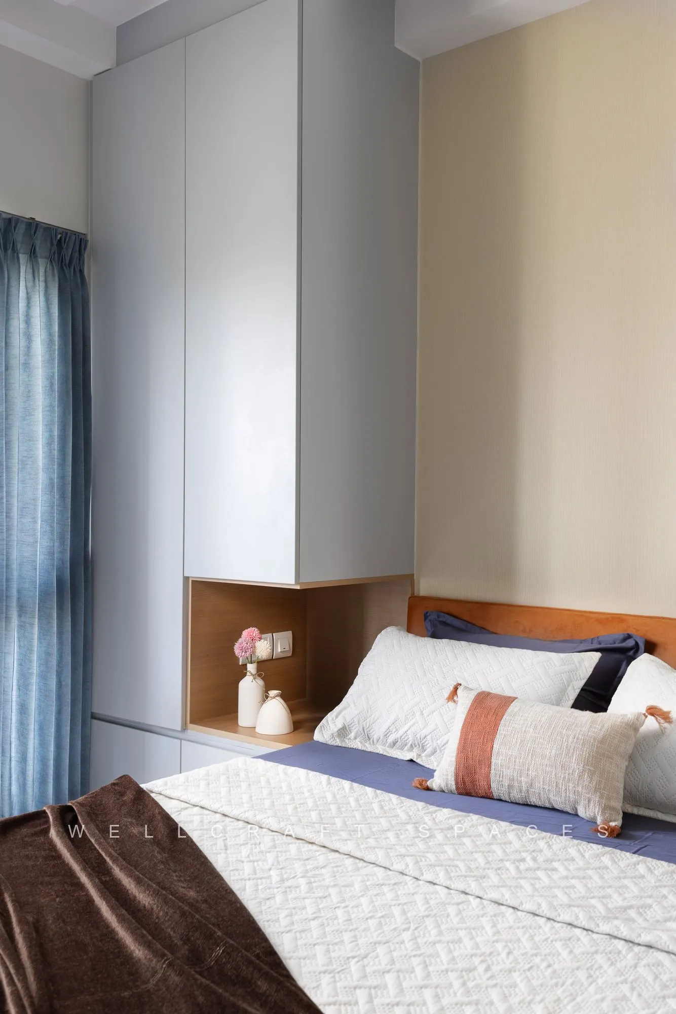 Guest bed with a walnut headboard and brass wall niche, dressed in white and brown linen beside blue sheer curtains.