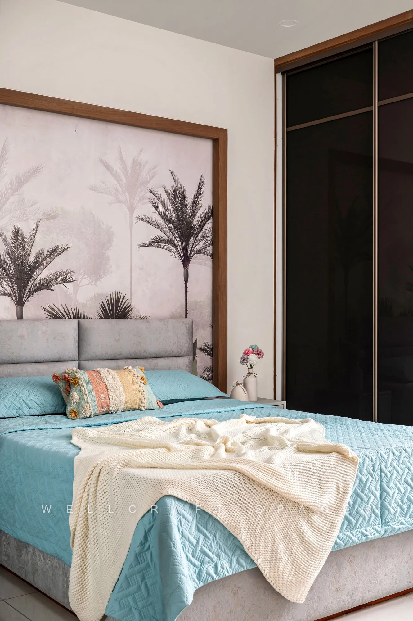 Bedroom with a painted palm mural behind a grey fabric headboard, teal quilted bedding with a cream throw and dark tinted glass wardrobe doors.